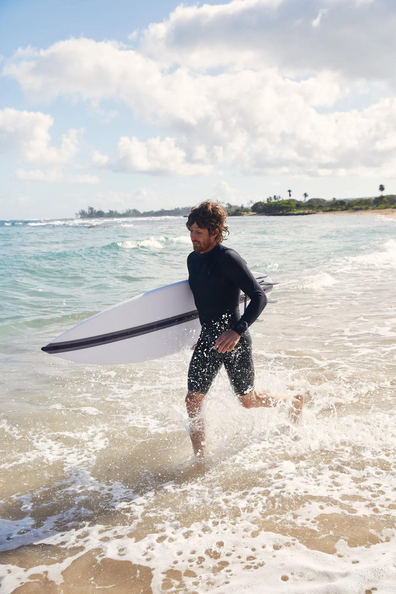 A man in a wetsuit carrying a surfboard while walking out of the ocean on a sunny day with clouds in the sky and a shoreline with trees in the background on the north shore of Oahu Hawaii.