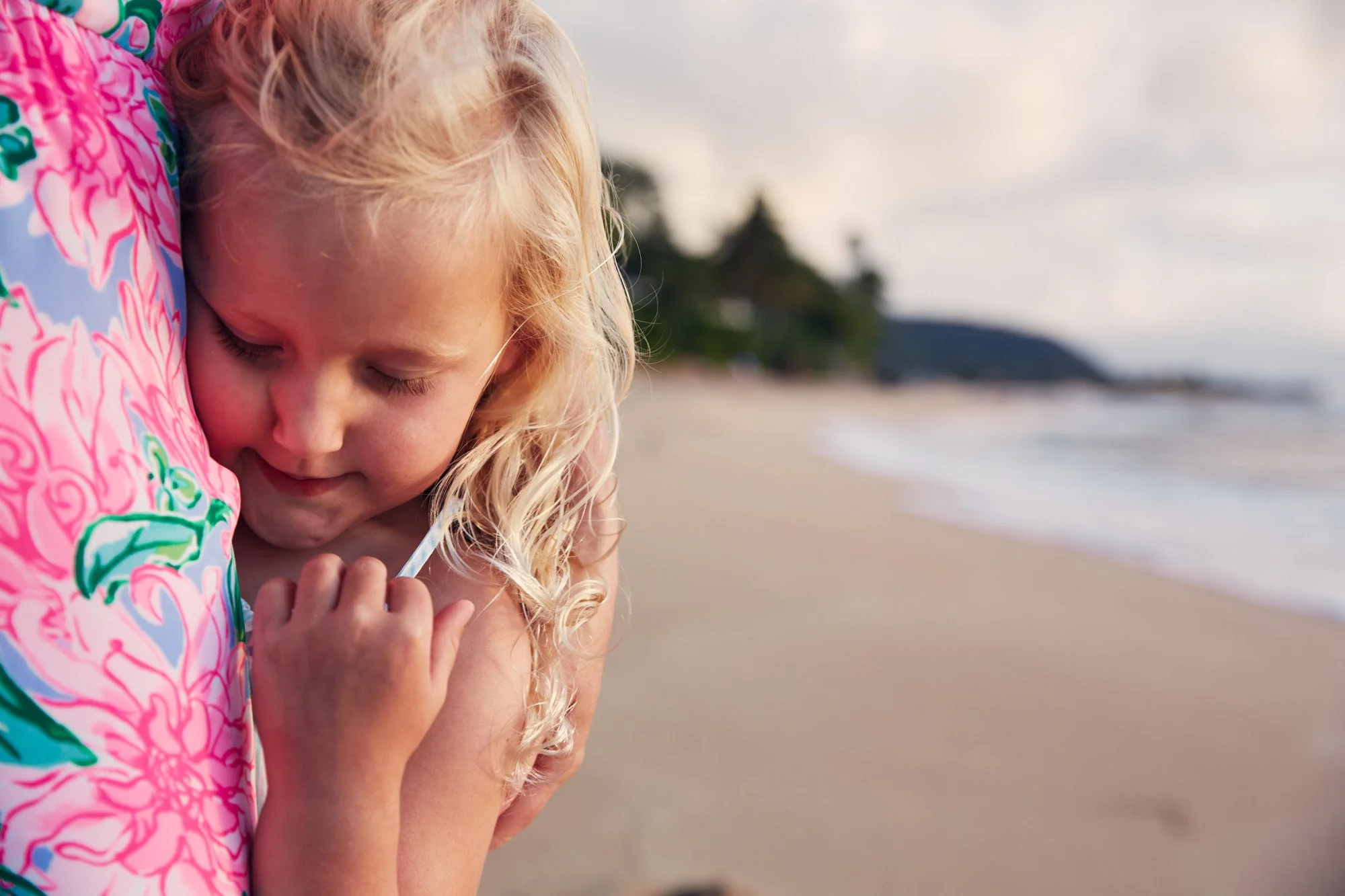 A young girl with blonde curly hair hugging her mother on a sandy beach on the north shore of Oahu Hawaii during a sunset with lit up clouds.