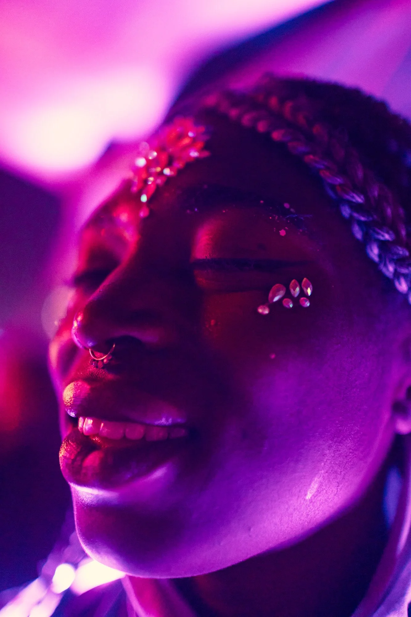 Close-up of a smiling person with dark skin, wearing a braided headband, with facial piercings, under colorful purple and pink lights. Burning Man