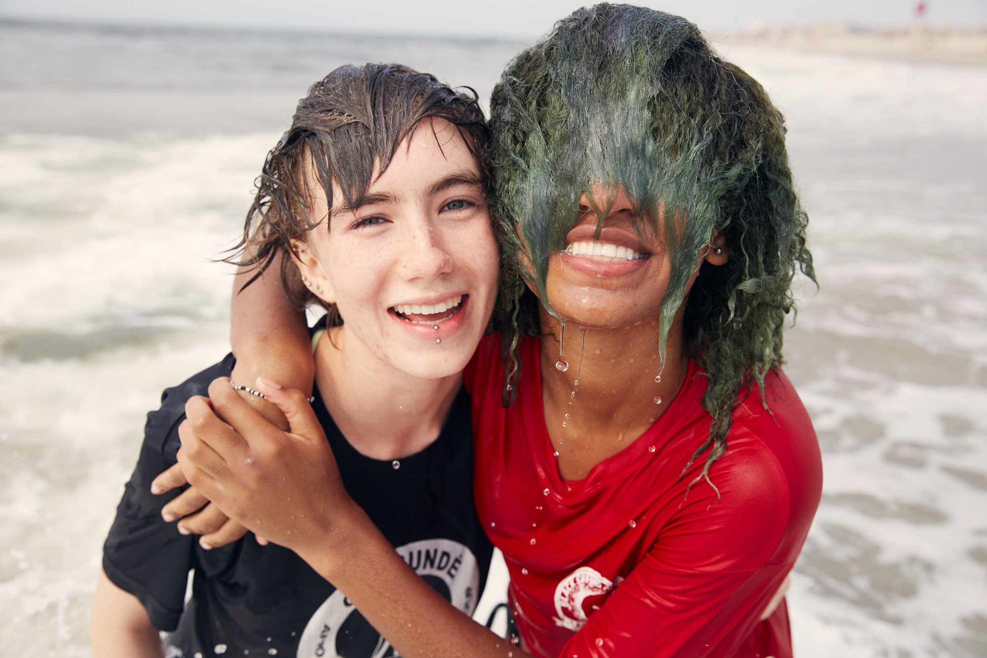 Two women hugging on the beach, joyful and smiling, with water dripping from their hair and faces.