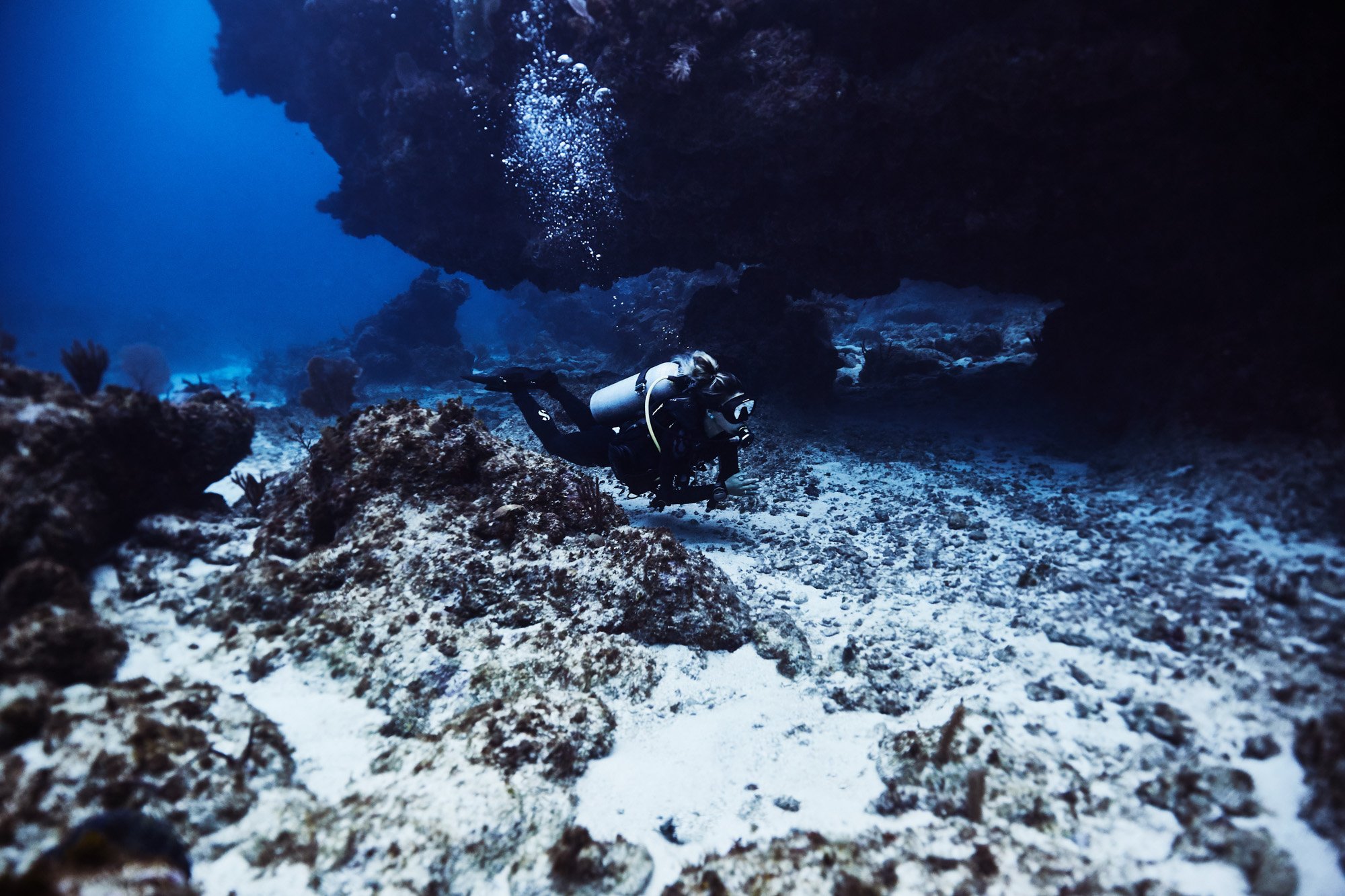 A scuba diver underwater exploring a rocky coral reef, with bubbles rising above, in Cayman Islands.