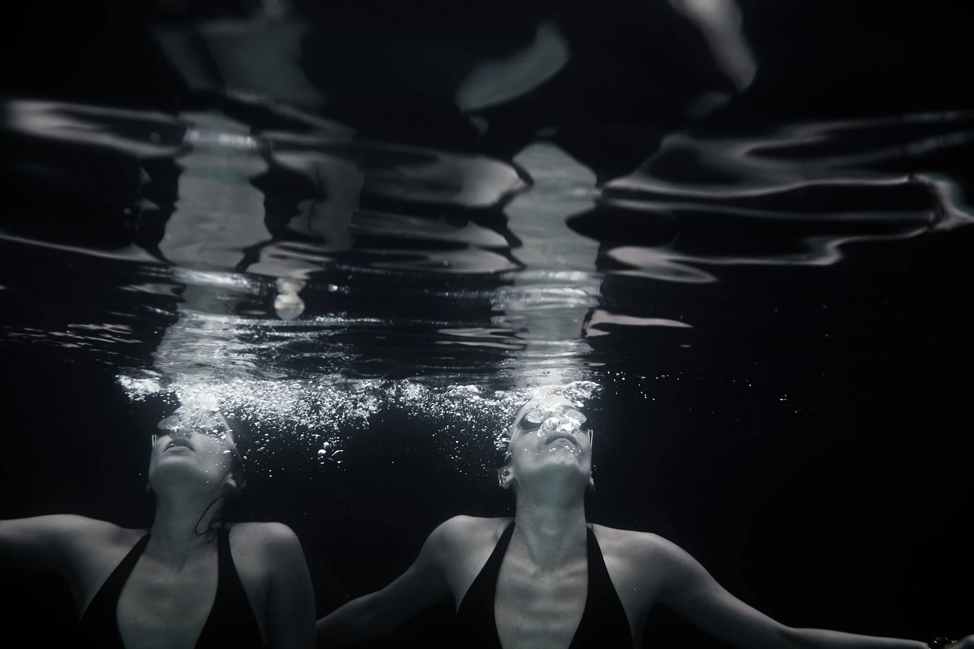 Two women Rachel and Rebekah Rife, underwater, wearing black swimsuits, floating with arms outstretched, with bubbles around their heads.