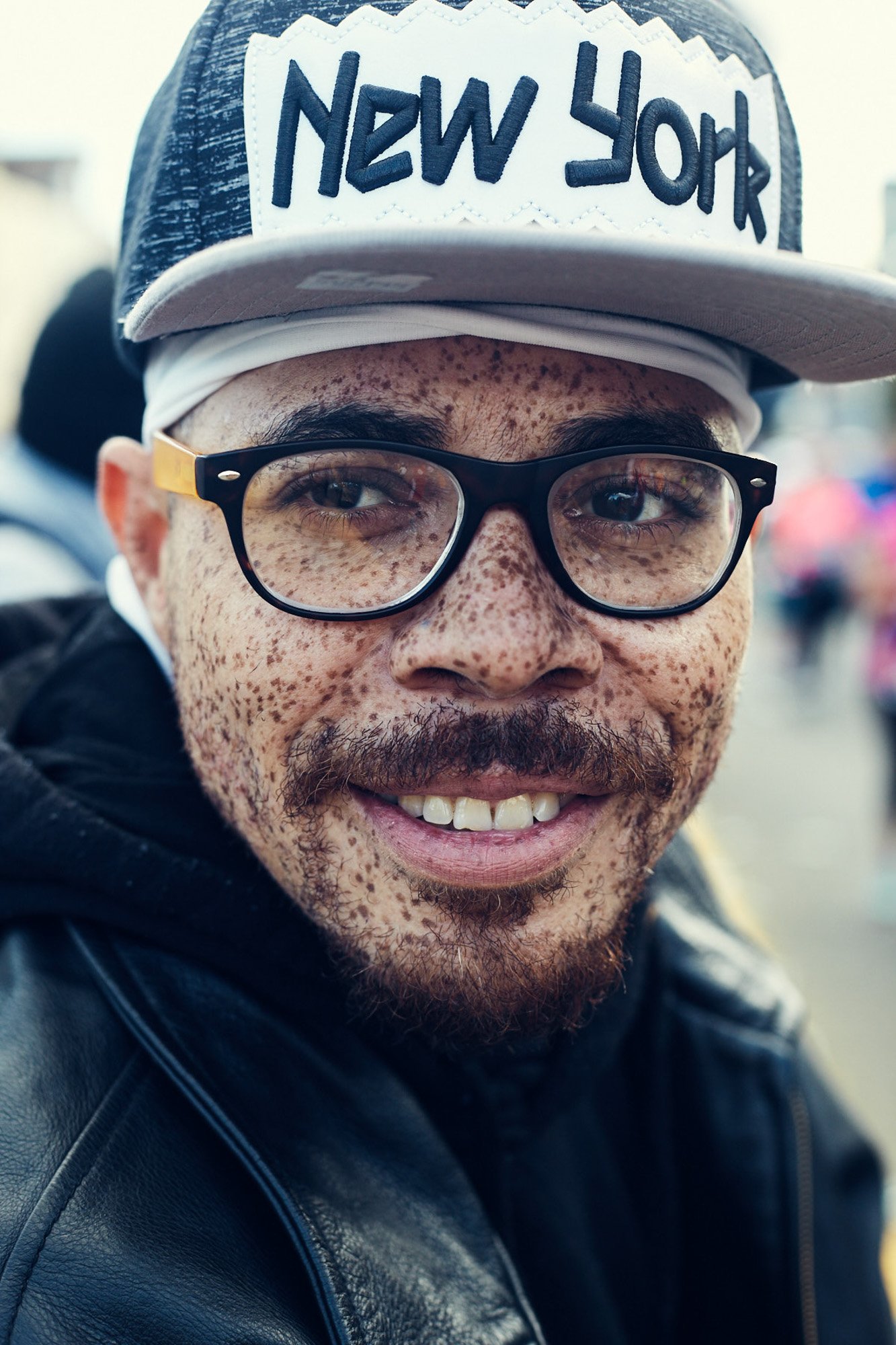 Close-up of a smiling man with glasses and freckles, wearing a gray hat with 'New York' written on it, and a black leather jacket, in a crowded outdoor setting.