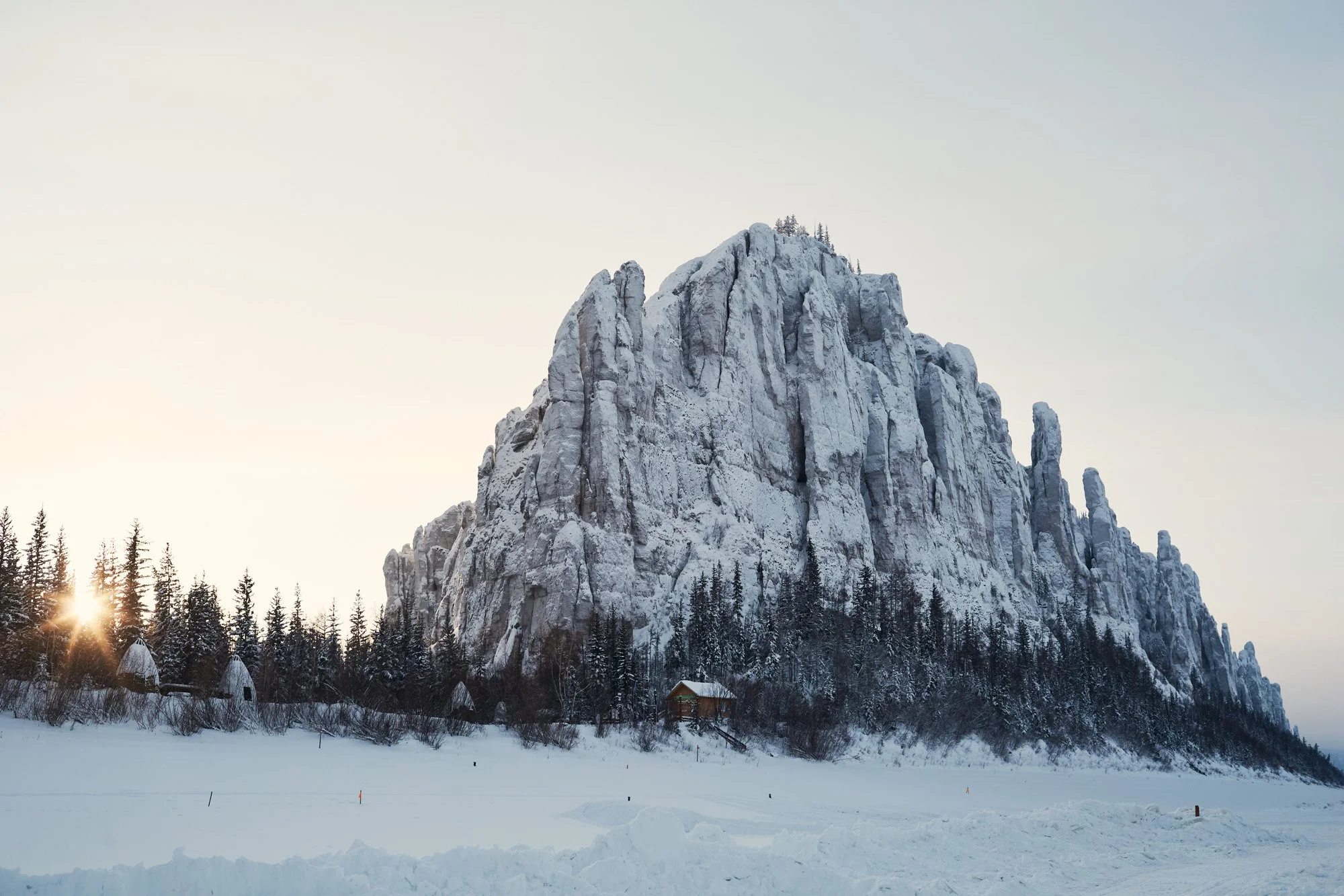A large snow-covered mountain with a forest at its base, Lena Pillars, in Sakha Republic near Yakutsk, Siberia Russia, small huts along the trees, and the sun setting behind the mountain in a winter landscape.