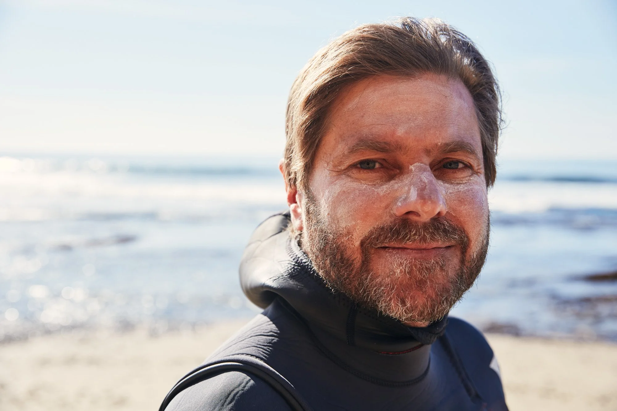 A man with a beard and mustache smiling at the camera on a beach near Scott's Creek near Santa Cruz California, wearing a black wetsuit and white sunscreen, with ocean and sky in the background.