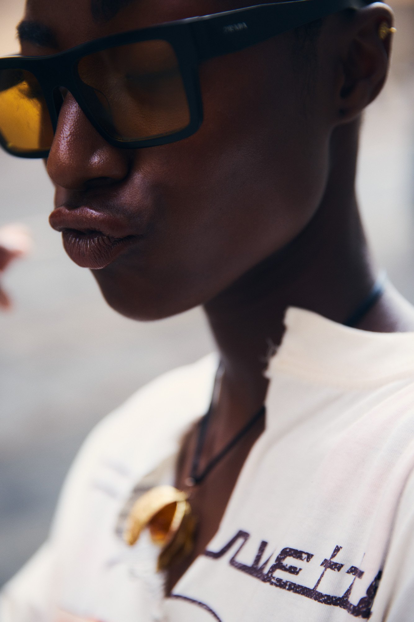 Close-up of a young Black woman wearing yellow-tinted sunglasses, a white shirt with black text, a black choker, and a gold pendant necklace.
