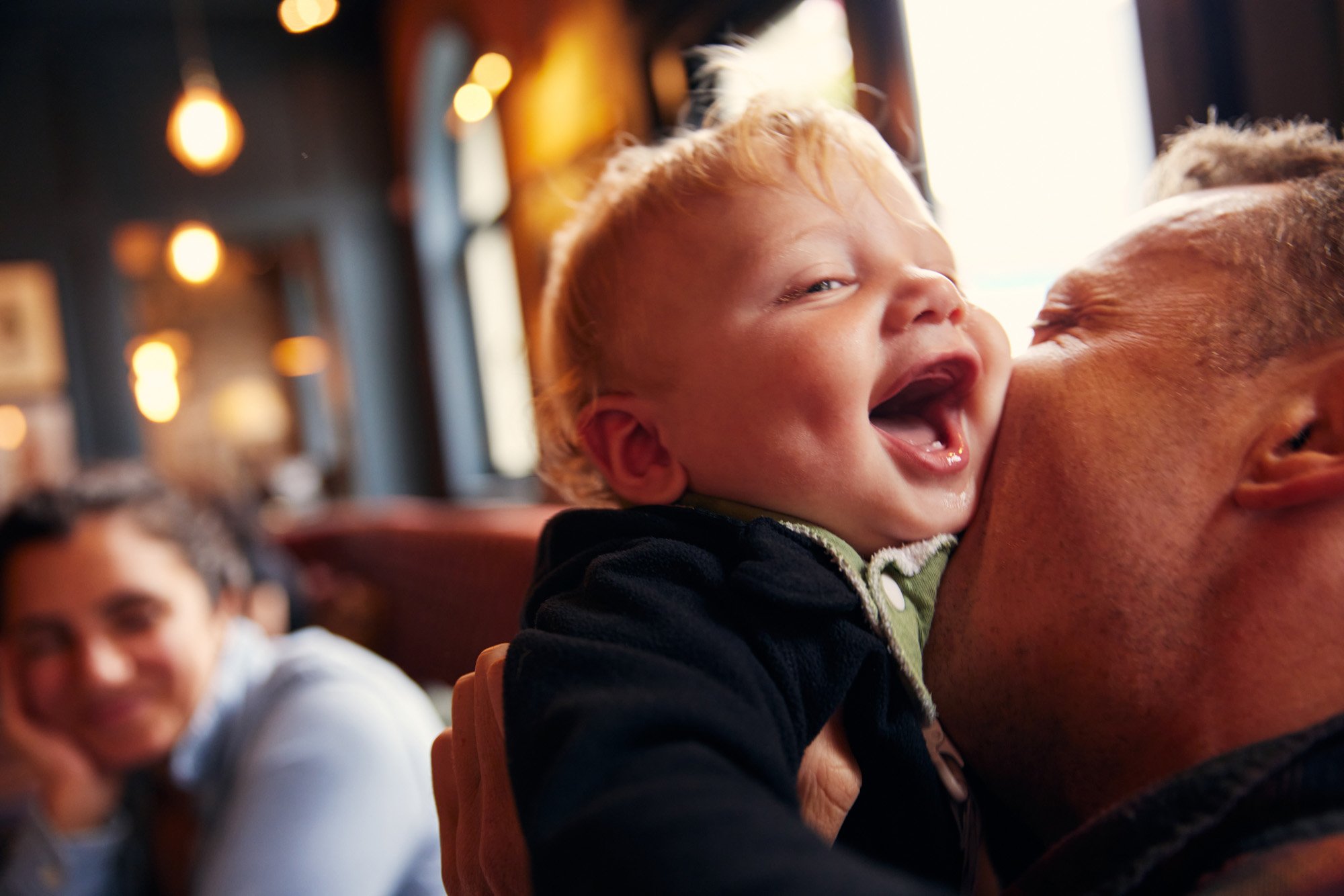 A toddler with curly blonde hair laughing and smiling while being held on an adult's shoulder in a cozy, warmly lit restaurant.