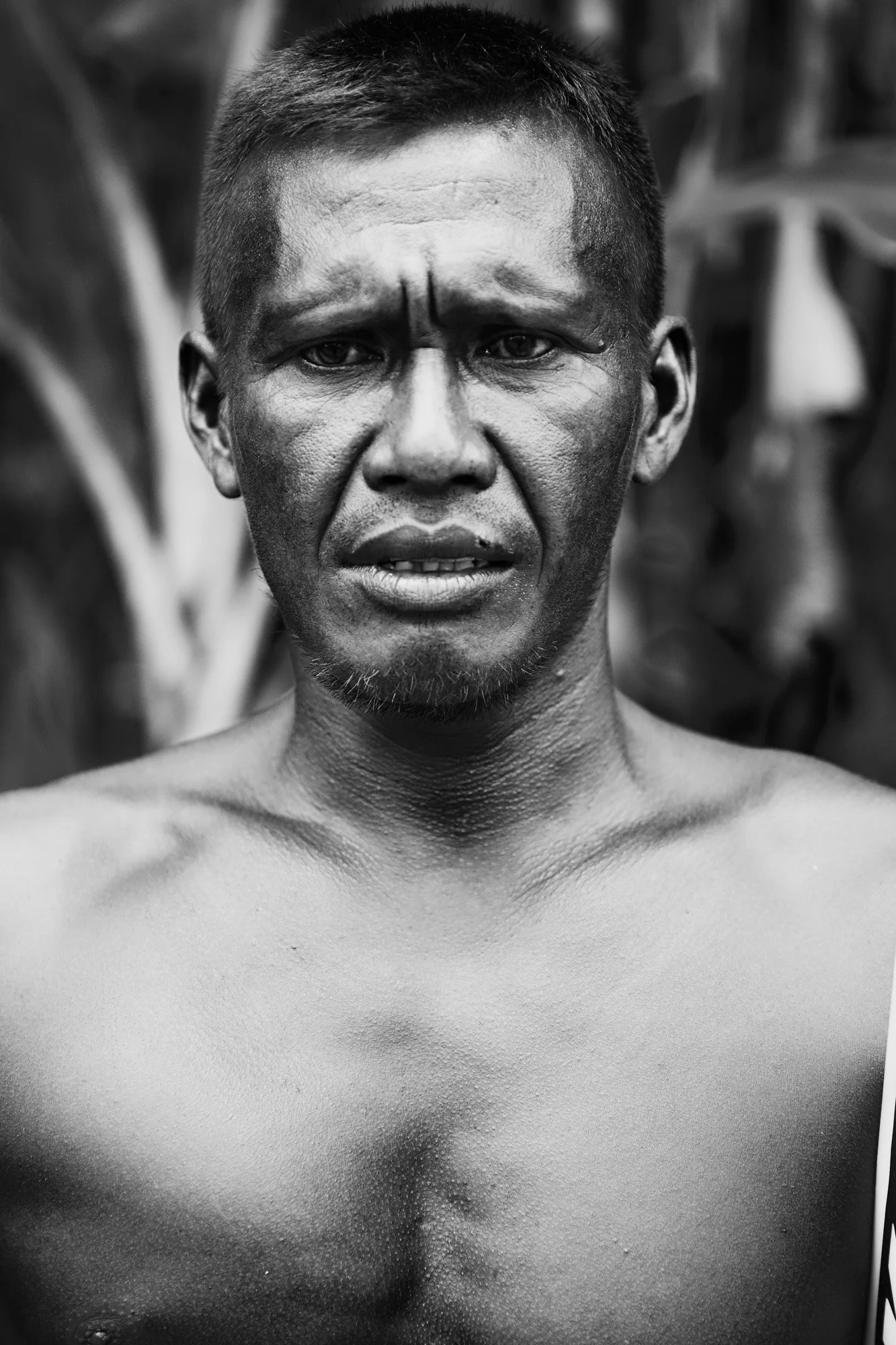 Black and white photo of a shirtless Balinese surfer with short hair and a serious expression, standing outdoors with blurred foliage in the background.