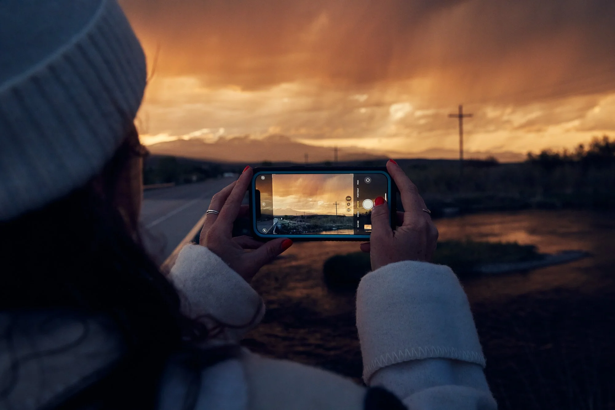 A woman taking a photo of a sunset with a smartphone on a rural road in Ennis Montana, with a cross and power lines in the distance.