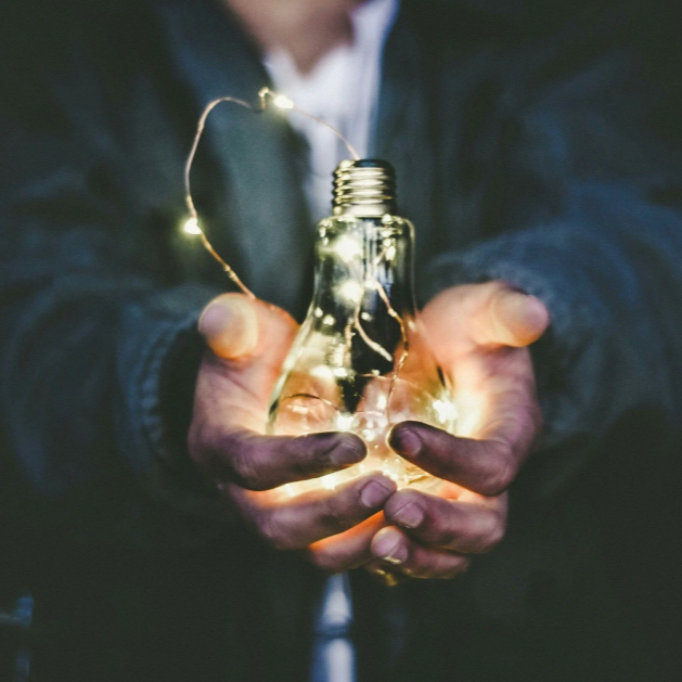 Person holding a light bulb filled with string lights, with a dark background.