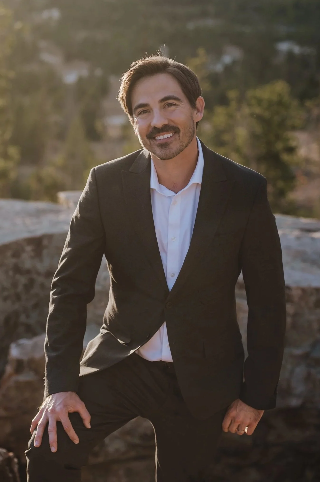 A man dressed in a black suit and white shirt, smiling outdoors with a blurred background of trees and rocks during sunset.