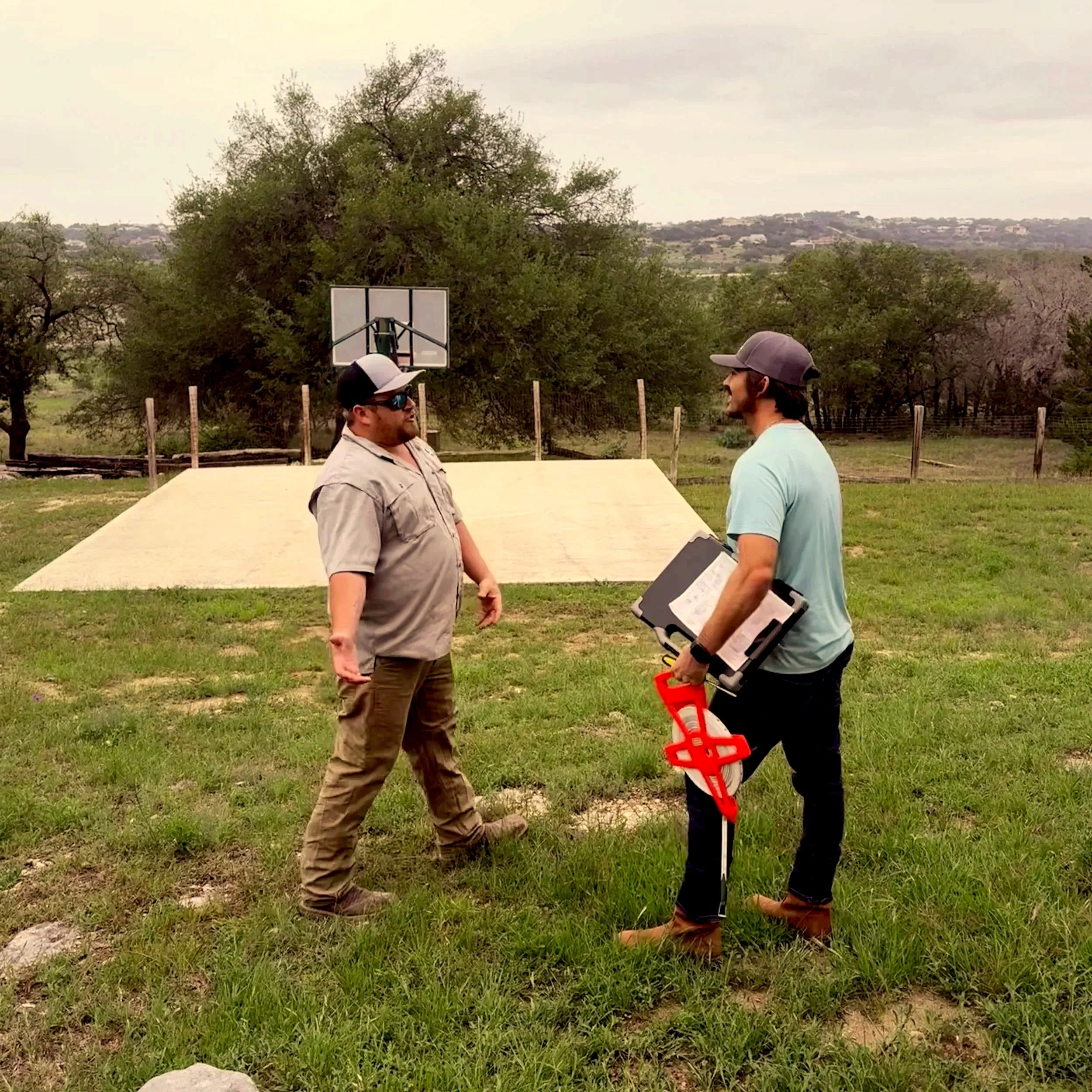 Two men standing outdoors on grass near a basketball hoop, engaged in a conversation. One is wearing sunglasses, a hat, and a beige shirt; the other is wearing a gray hat, a light blue shirt, and holding a tool with a red handle.