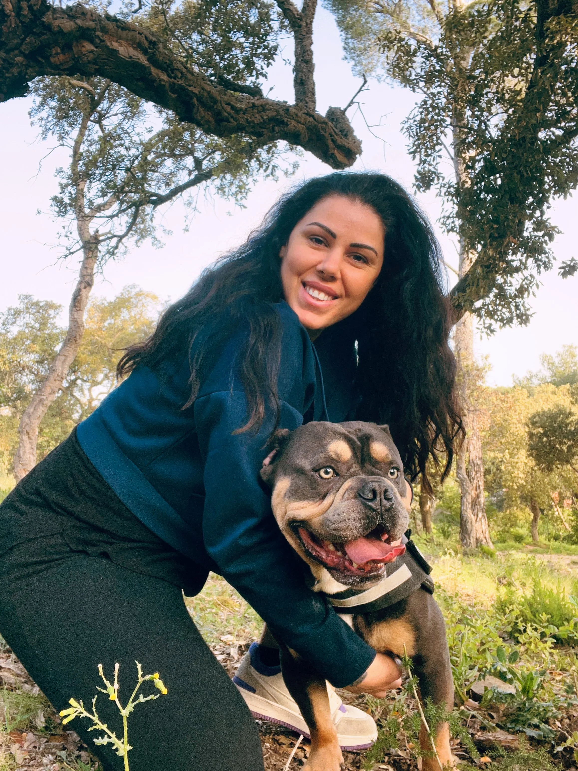 Une femme souriante à côté d'un chien Pitbull dans un environnement naturel avec des arbres.