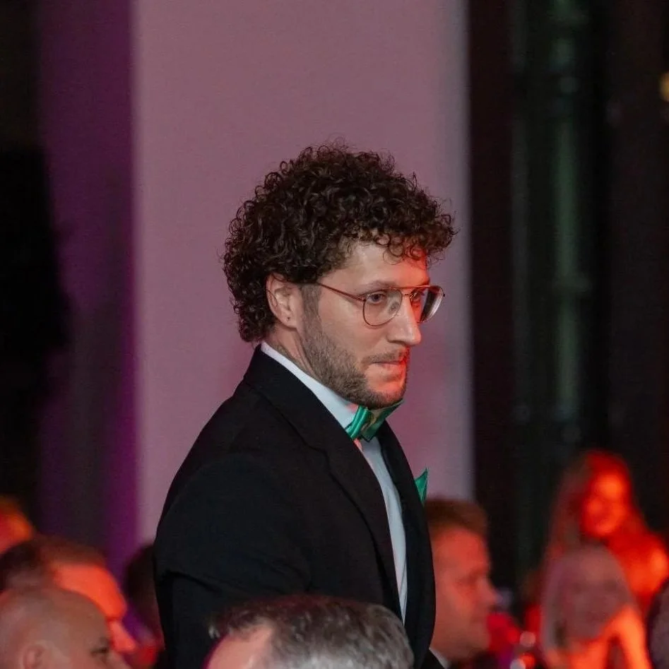 Ein Mann mit Brille und lockigem Haar trägt einen schwarzen Anzug, weiße Hemd und grüne Fliege auf einer formellen Veranstaltung.