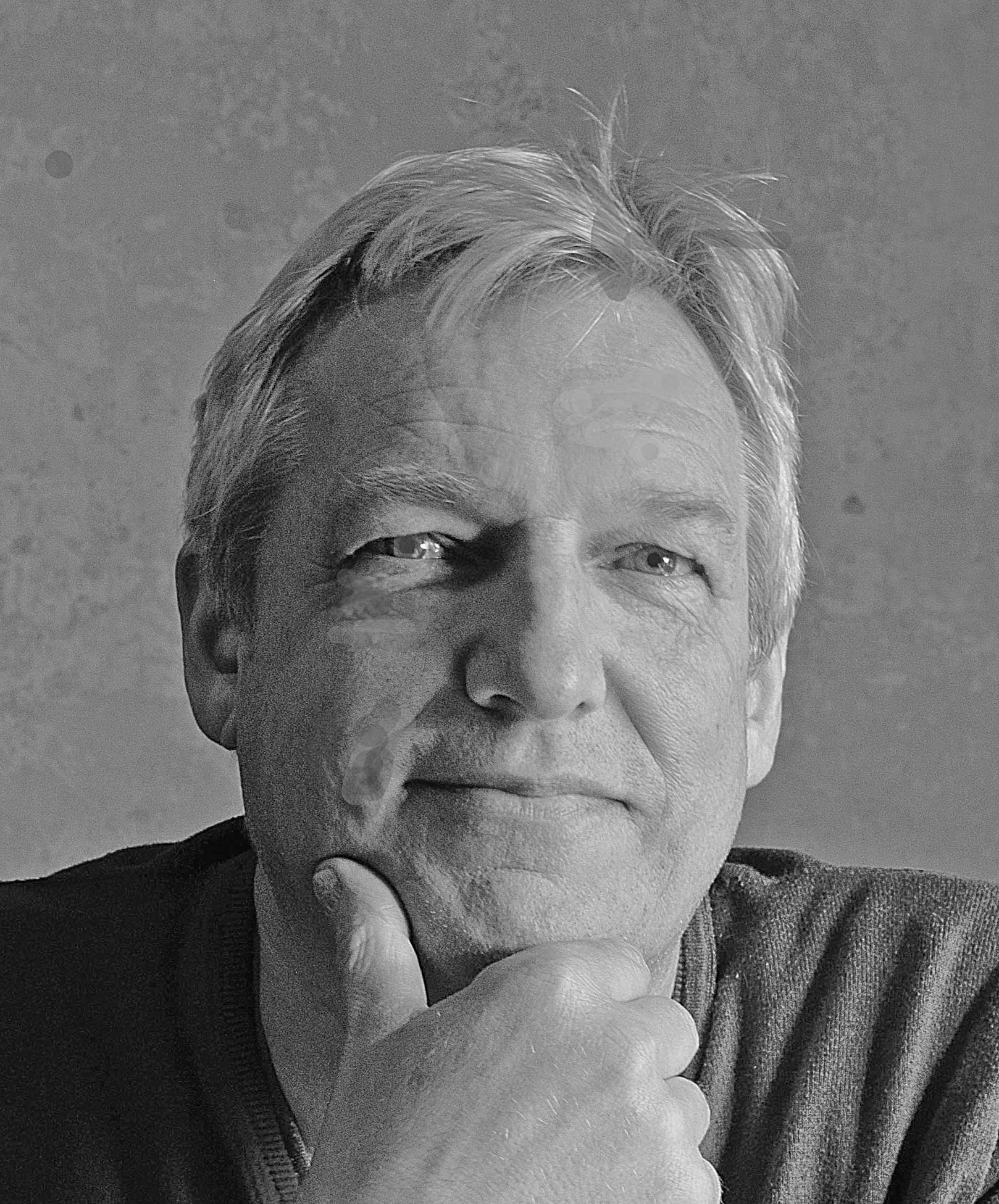 Un portrait d'un homme aux cheveux gris, avec une expression pensante, se tenant le menton avec la main, en noir et blanc.