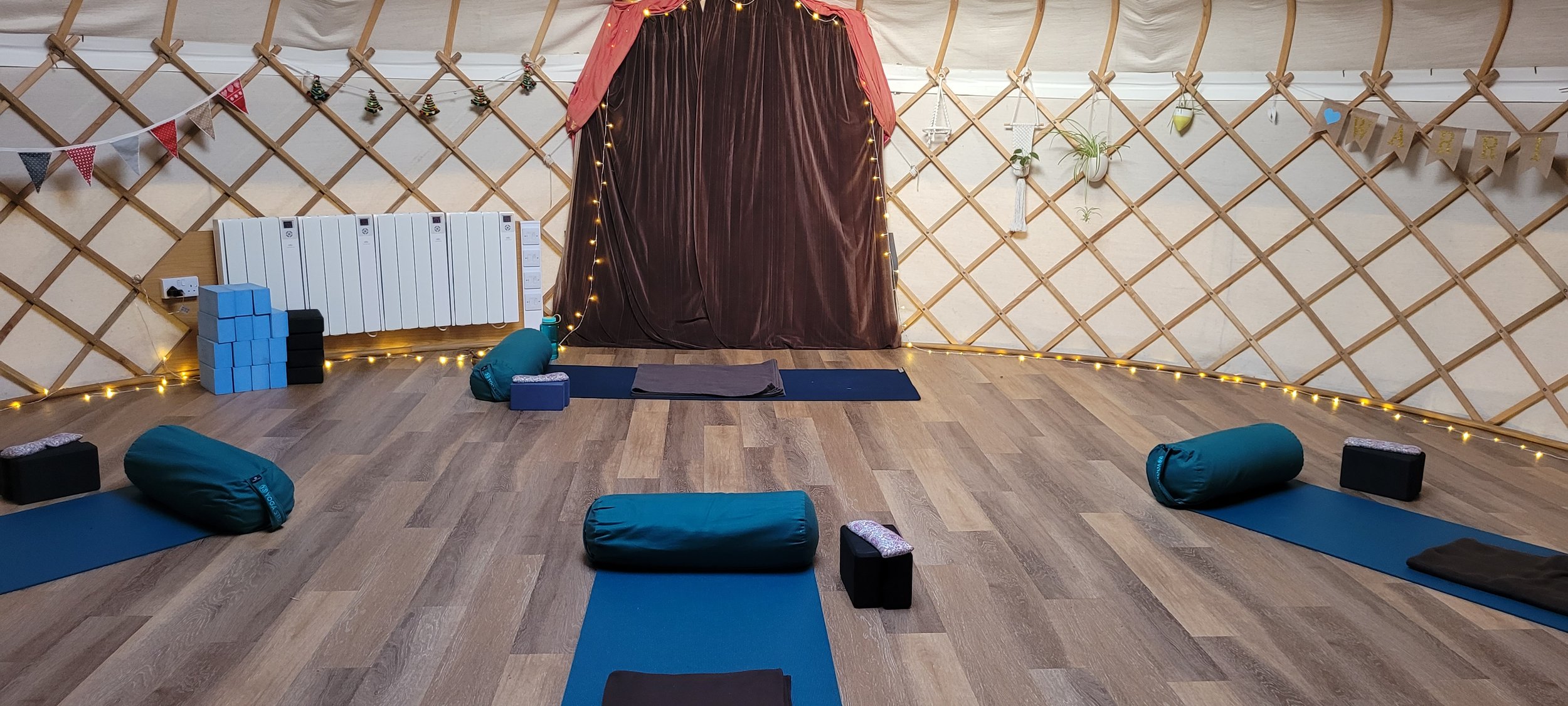 A cozy indoor yoga or meditation space with yoga mats, bolsters, and blocks arranged on a wooden floor. The background features a lattice wall decorated with string lights, small hanging plants, and a pink curtain framing a dark curtain or backdrop.