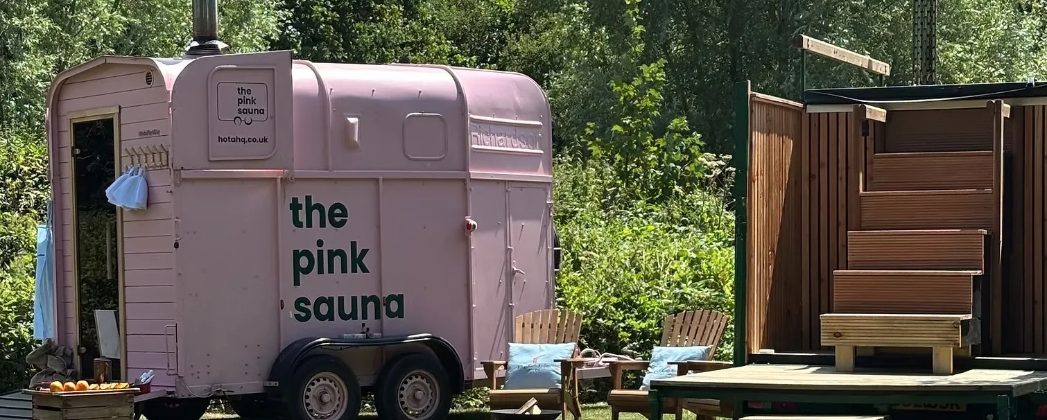 Pink mobile sauna trailer labeled 'the pink sauna' and 'thepinksauna.co.uk,' with two outdoor wooden chairs with cushions and wooden stairs leading to a small outdoor shower or changing area, set in a lush green garden.