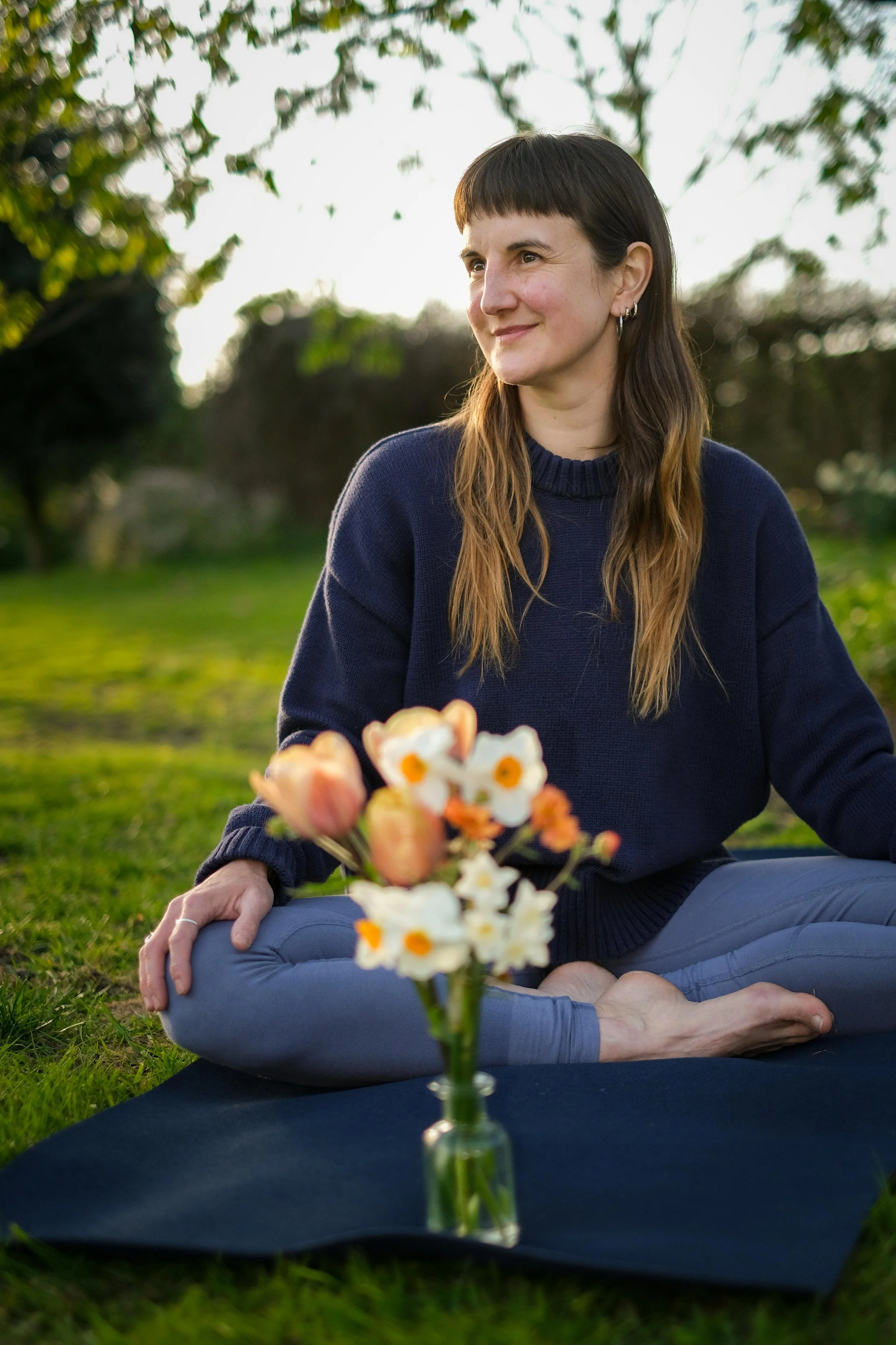 A woman with brown hair and bangs, wearing a navy blue sweater, sitting outdoors in a meditative pose on the grass, with a bouquet of flowers in front of her and trees in the background.