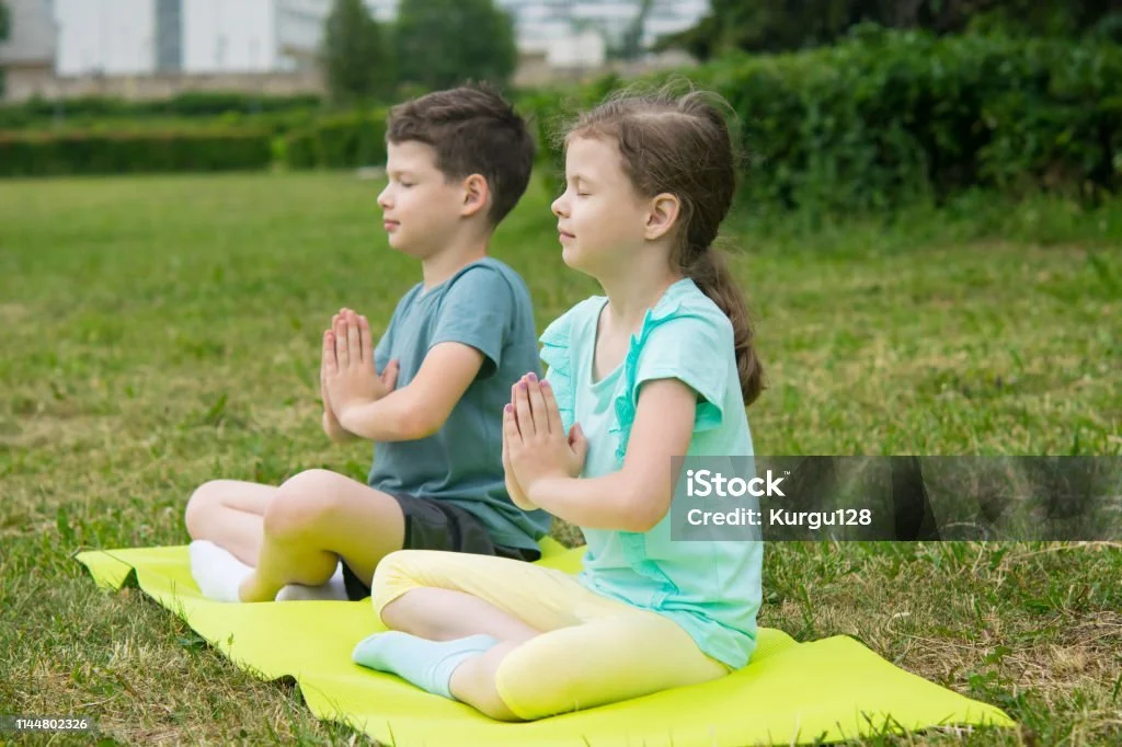 Two children, a boy and a girl, sitting cross-legged on a yoga mat outdoors with their eyes closed, practicing meditation or mindfulness.