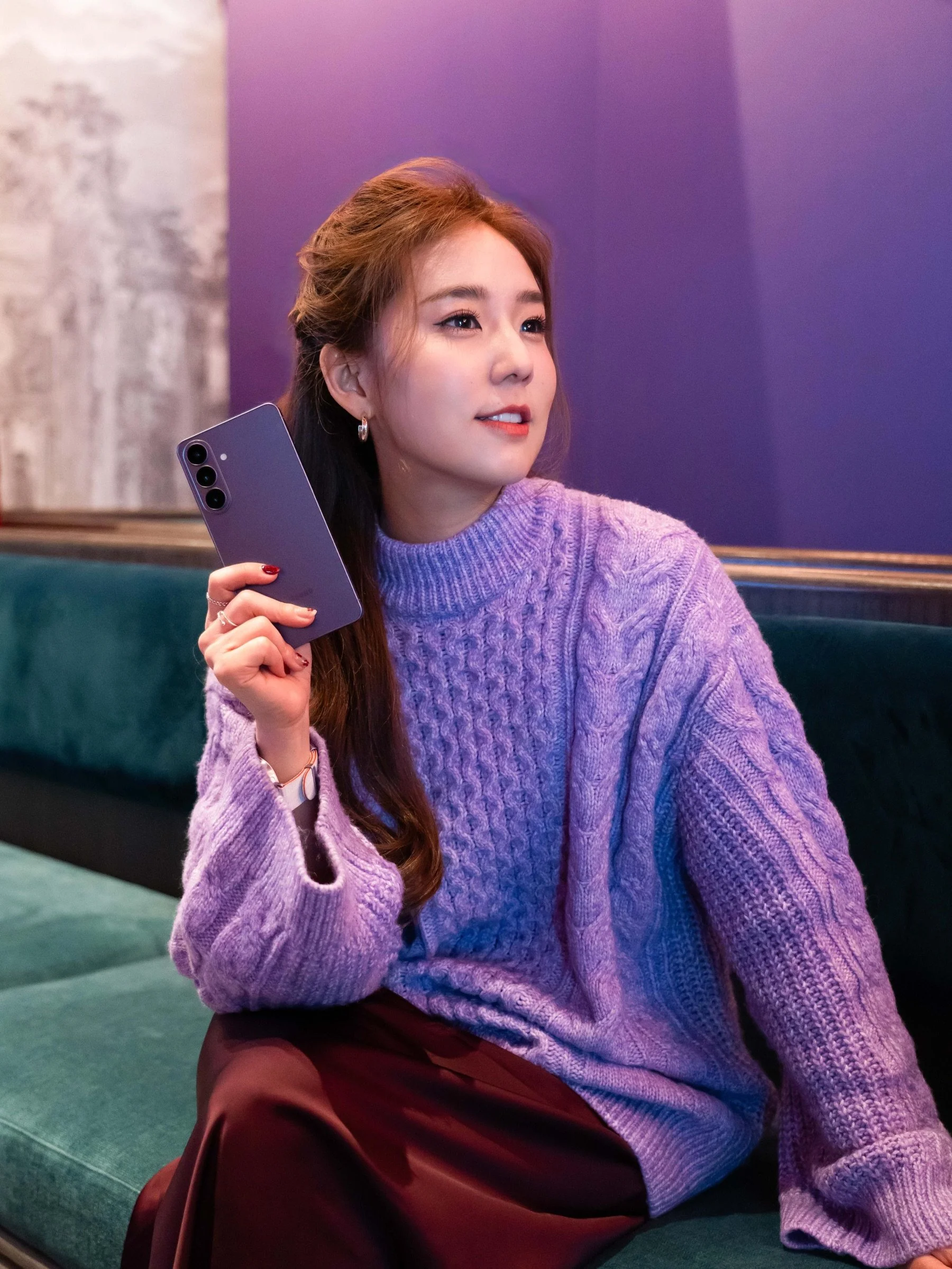 A young woman with long brown hair wearing a lavender cable-knit sweater and maroon skirt, sitting on a green couch against a purple wall, holding a purple smartphone near her face. Make up by make up artist Muacher (Cheron Tan)