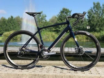 A black bicycle with a blue frame parked on a paved area near a body of water, with trees and a fountain in the background.