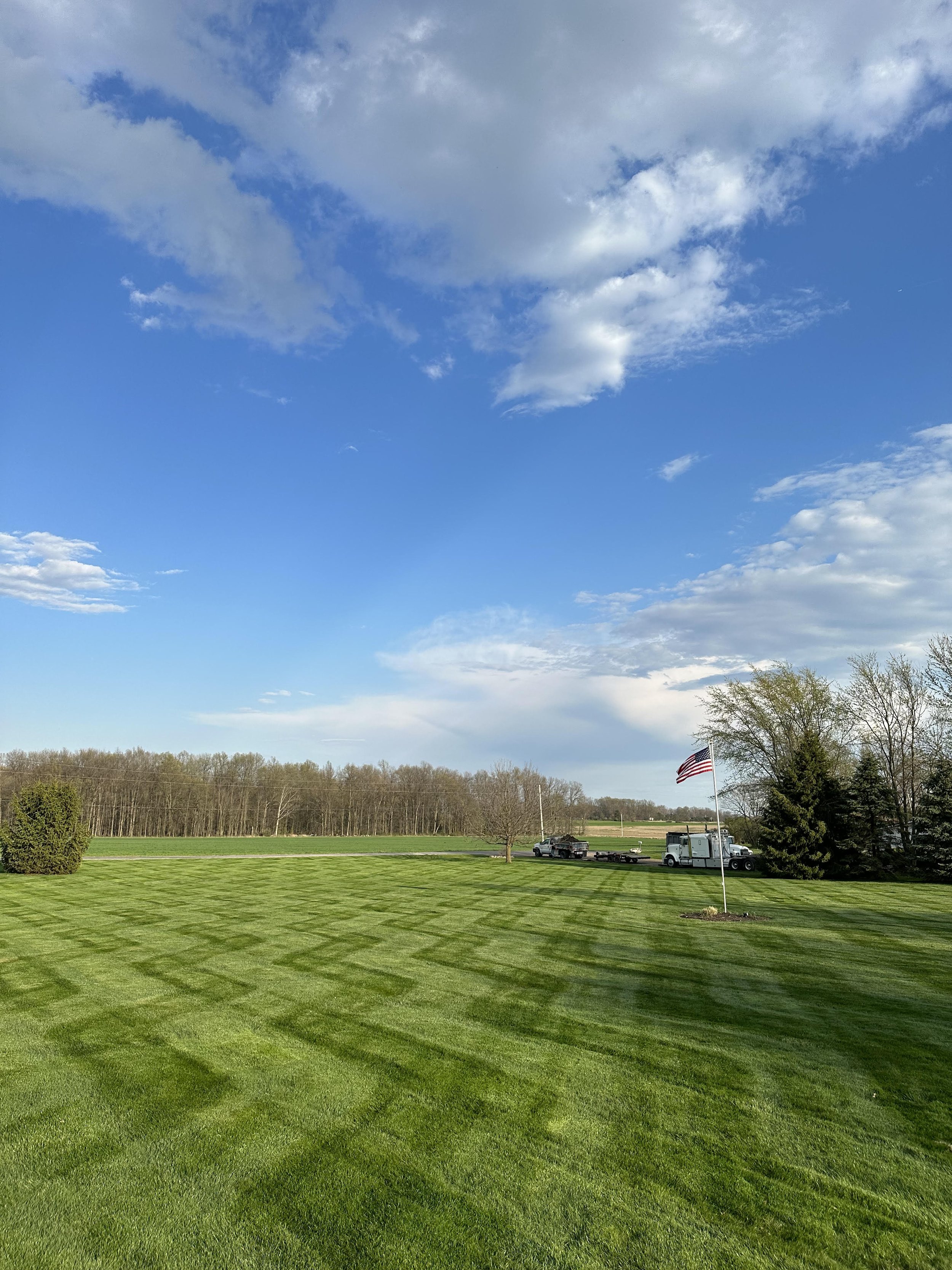 A well-manicured green lawn with a flagpole flying an American flag on a sunny day. There are trees and a truck in the background under a partly cloudy sky.