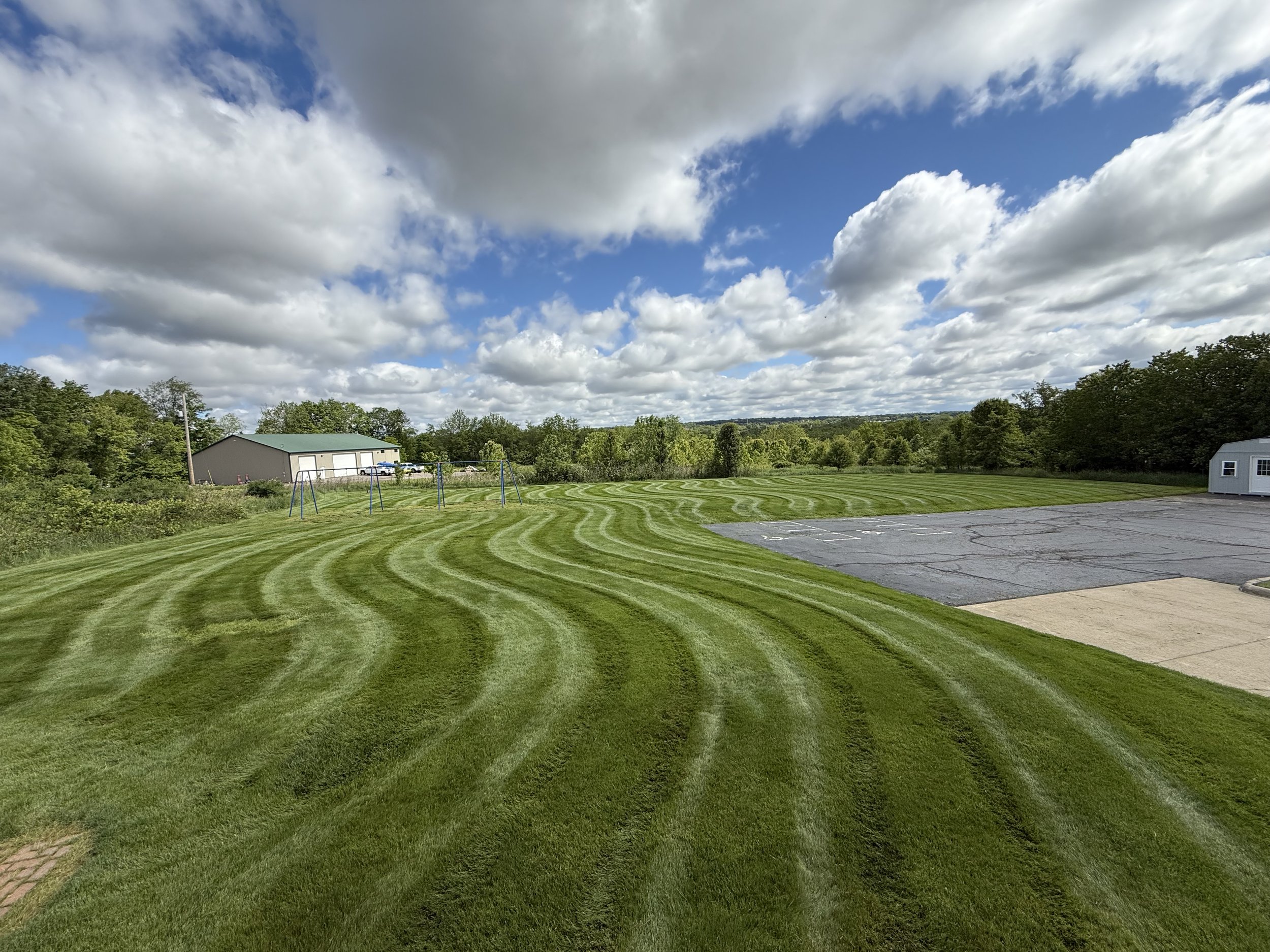 A well-manicured grassy field with curved mowing patterns, a few playground swings, a parking lot with some cars, a small building, and a background of trees under a partly cloudy sky.