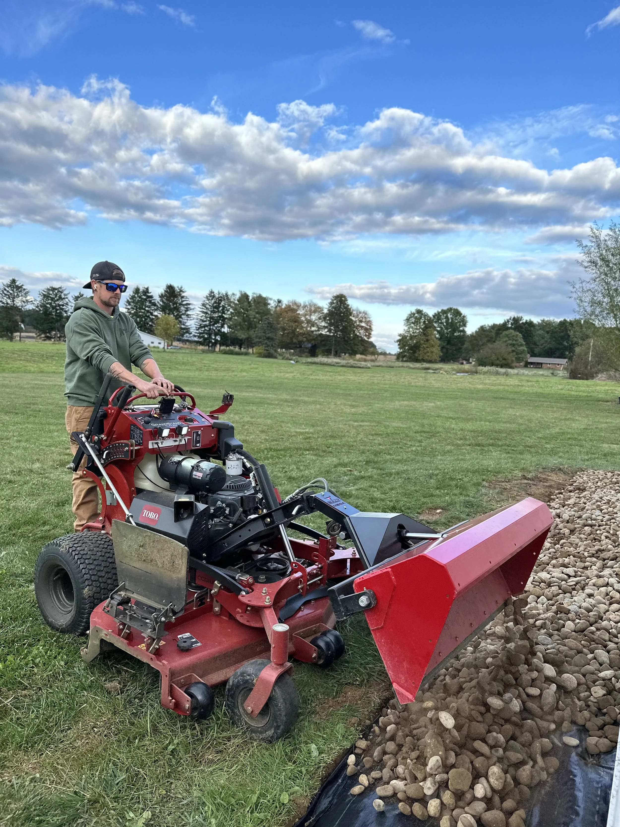 A man operating a red and black soil debris remover machine on a grassy field with trees and a partly cloudy sky in the background.