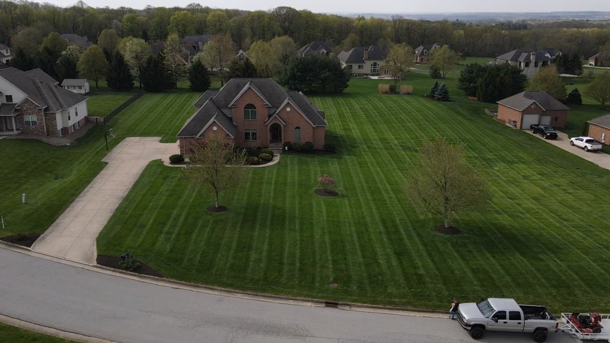 An aerial view of a suburban neighborhood with a large brick house, a well-maintained lawn, a driveway, and a white pickup truck parked on the street.