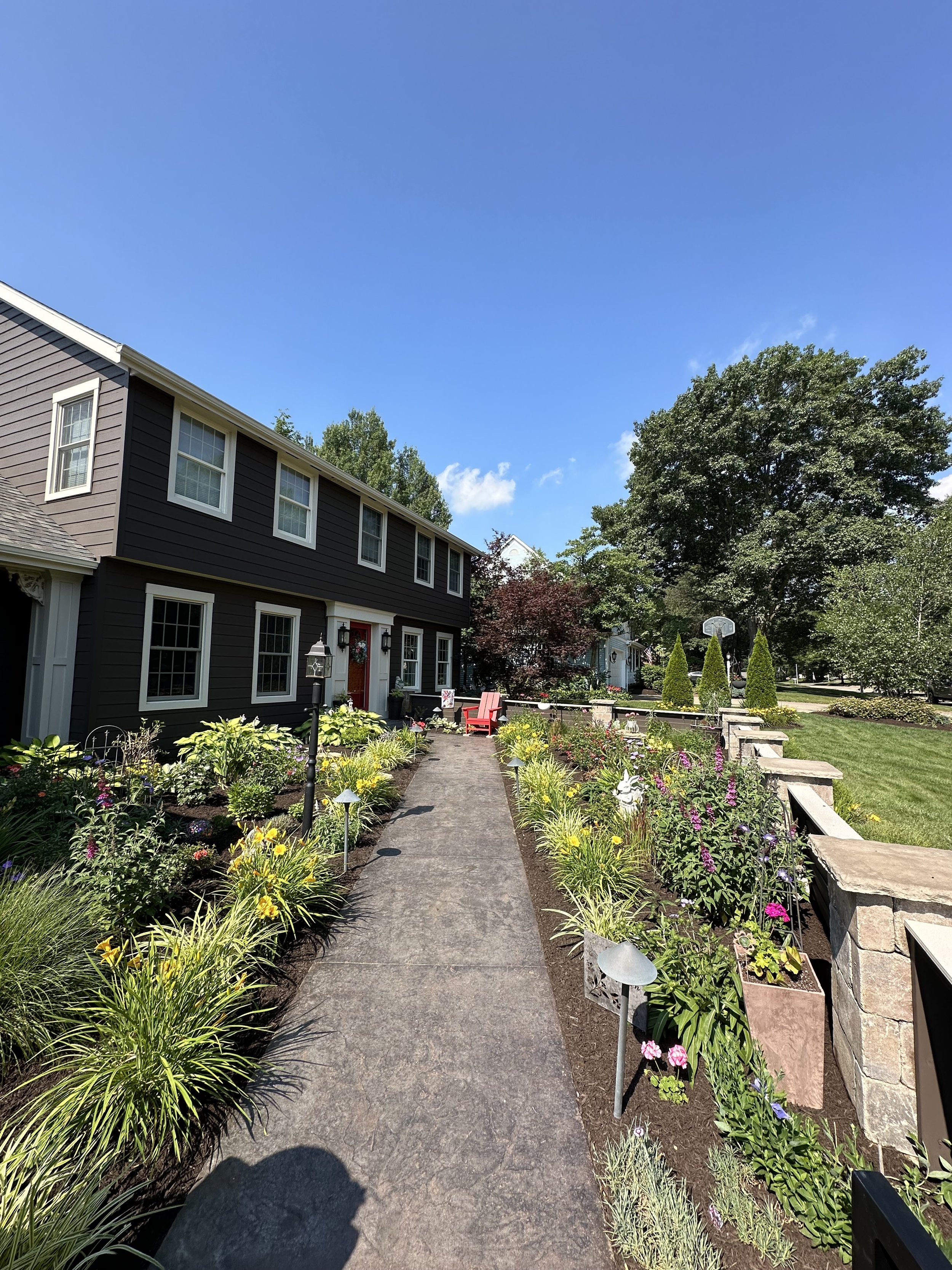 A residential house with a garden path, colorful flowers, and outdoor furniture under a clear blue sky.