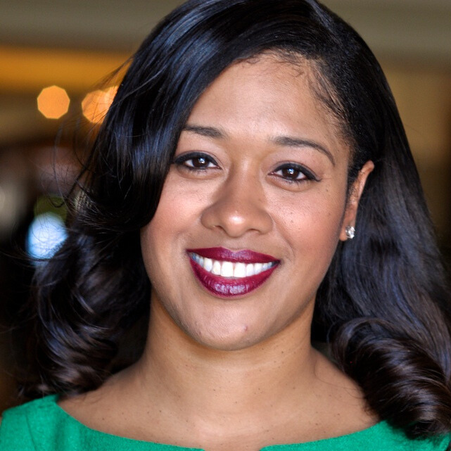 Close-up portrait of a woman with dark hair, wearing a green top, smiling with dark lipstick, in an indoor setting.