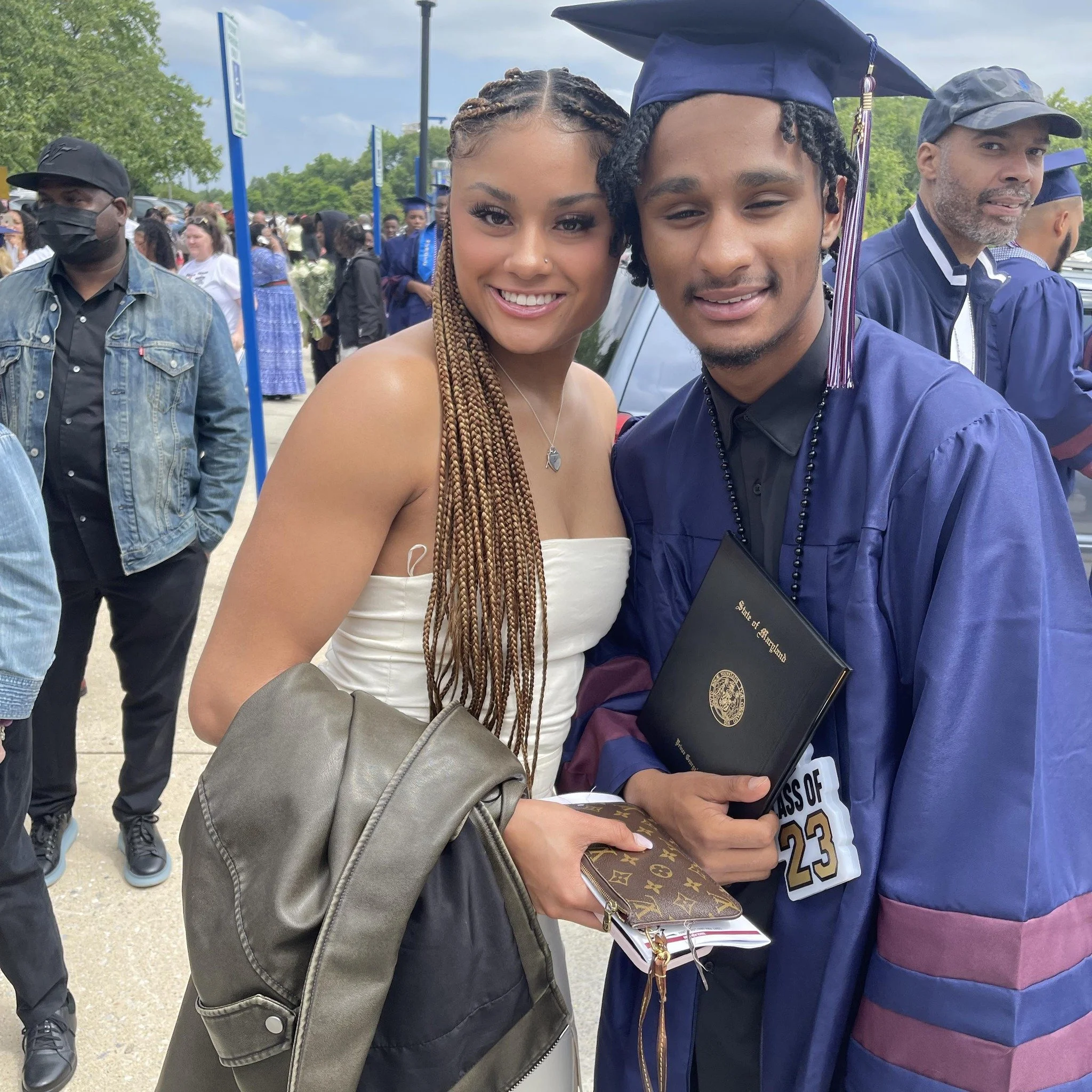A young man in a graduation cap and gown holding a diploma, standing next to a young woman in a white strapless dress, both smiling at a graduation ceremony outdoors.
