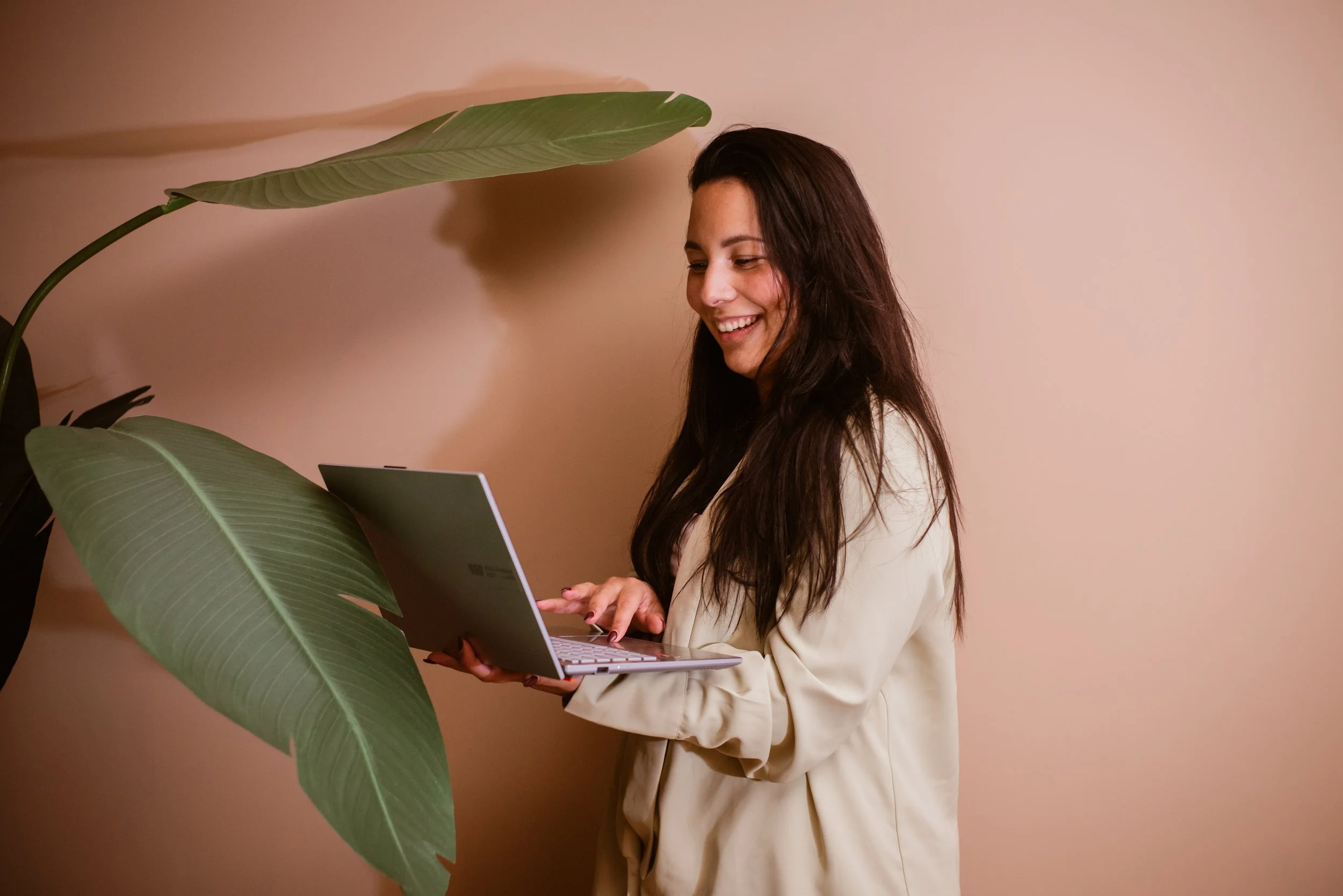 Woman with long dark hair in a beige blazer using a laptop near a large green plant with big leaves against a beige wall.
