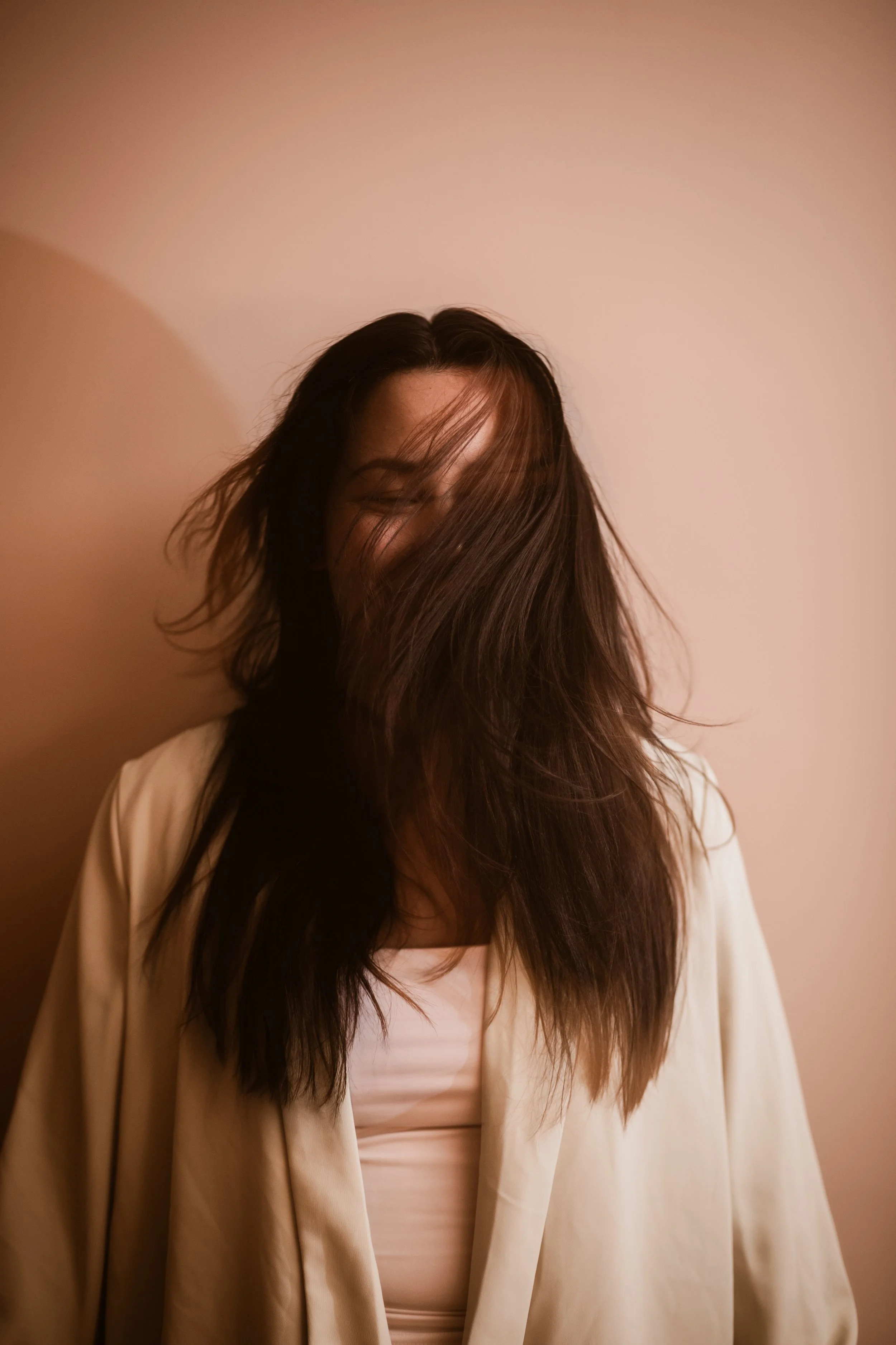 A woman with long, dark hair partially covering her face, standing against a plain background, wearing a light-colored blazer over a white top.
