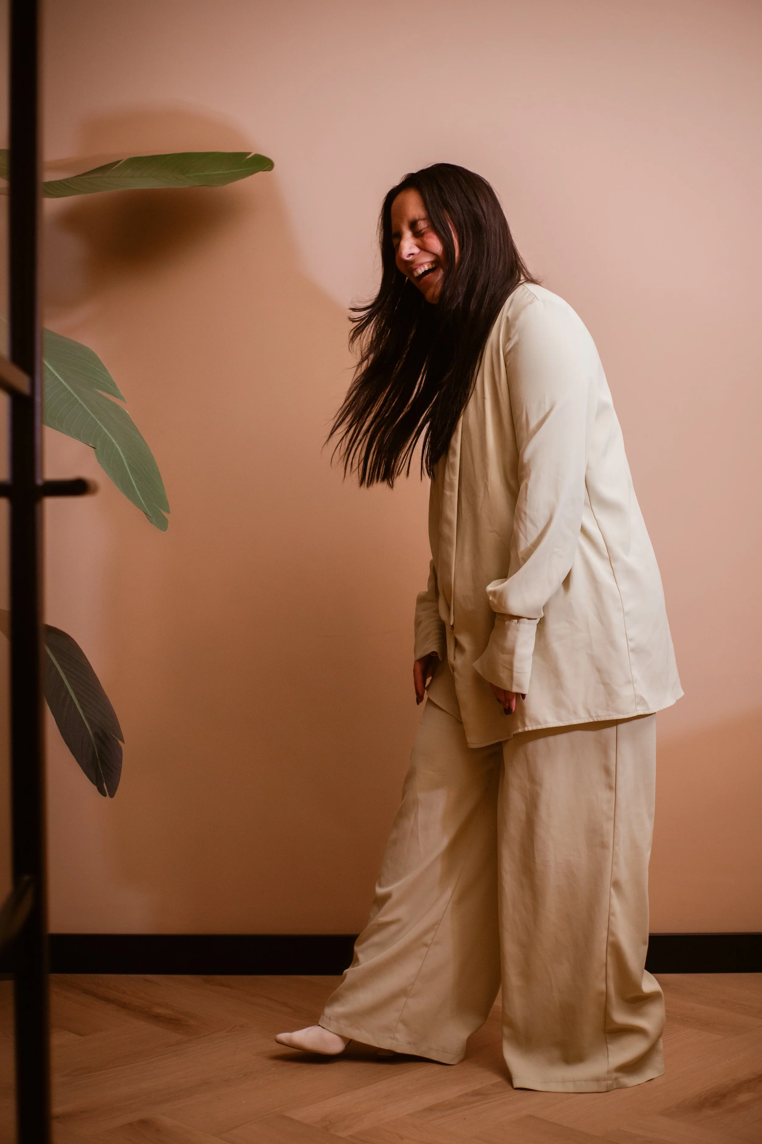 A woman in cream-colored pajamas laughing and leaning forward against a beige wall with wooden flooring, partially obscured by a green potted plant.