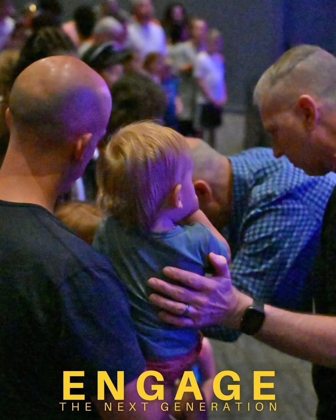 A group of men, including one holding a young child, engaged in a serious conversation in a crowded indoor setting with many people in the background.