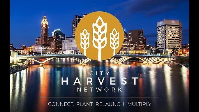 Nighttime city skyline with illuminated buildings and reflection on river, overlay with City Harvest Network logo featuring wheat icons and text.