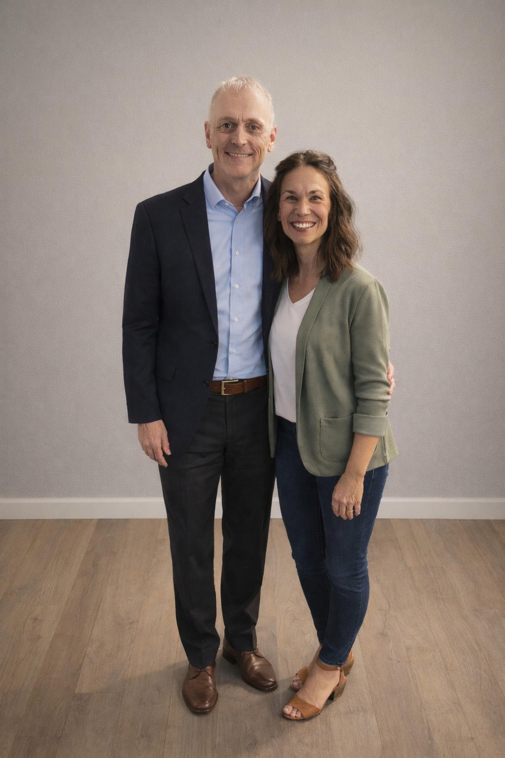 A man and woman smiling, standing close together in front of a plain wall. The man is tall with gray hair, wearing a dark suit, light blue shirt, and brown shoes. The woman has shoulder-length brown hair, wearing a green blazer, white shirt, jeans, and tan heels.