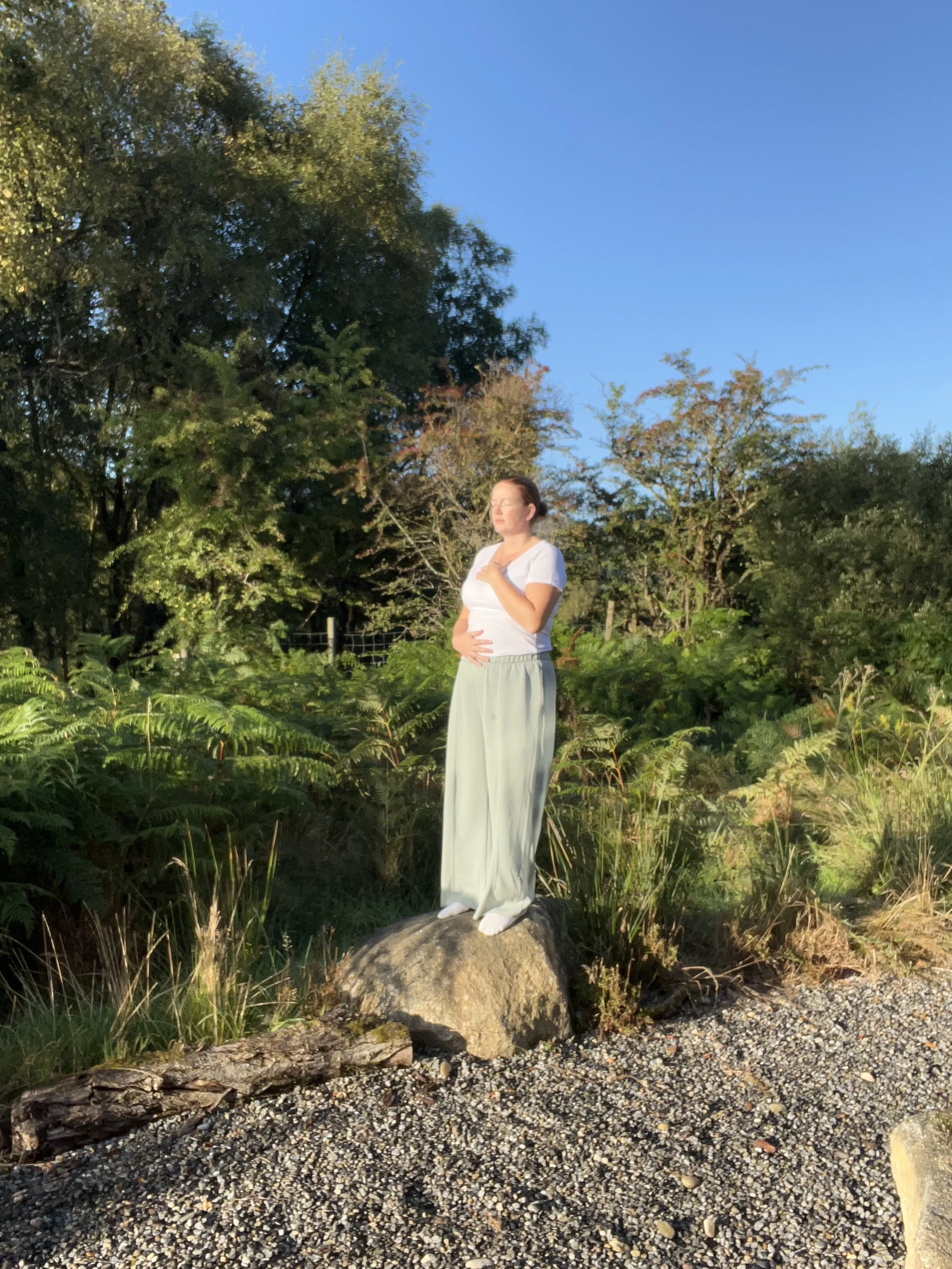 A woman standing on a large rock in a natural outdoor setting, eyes closed, with sun shining on her face and belly, surrounded by greenery and trees, under a clear blue sky.