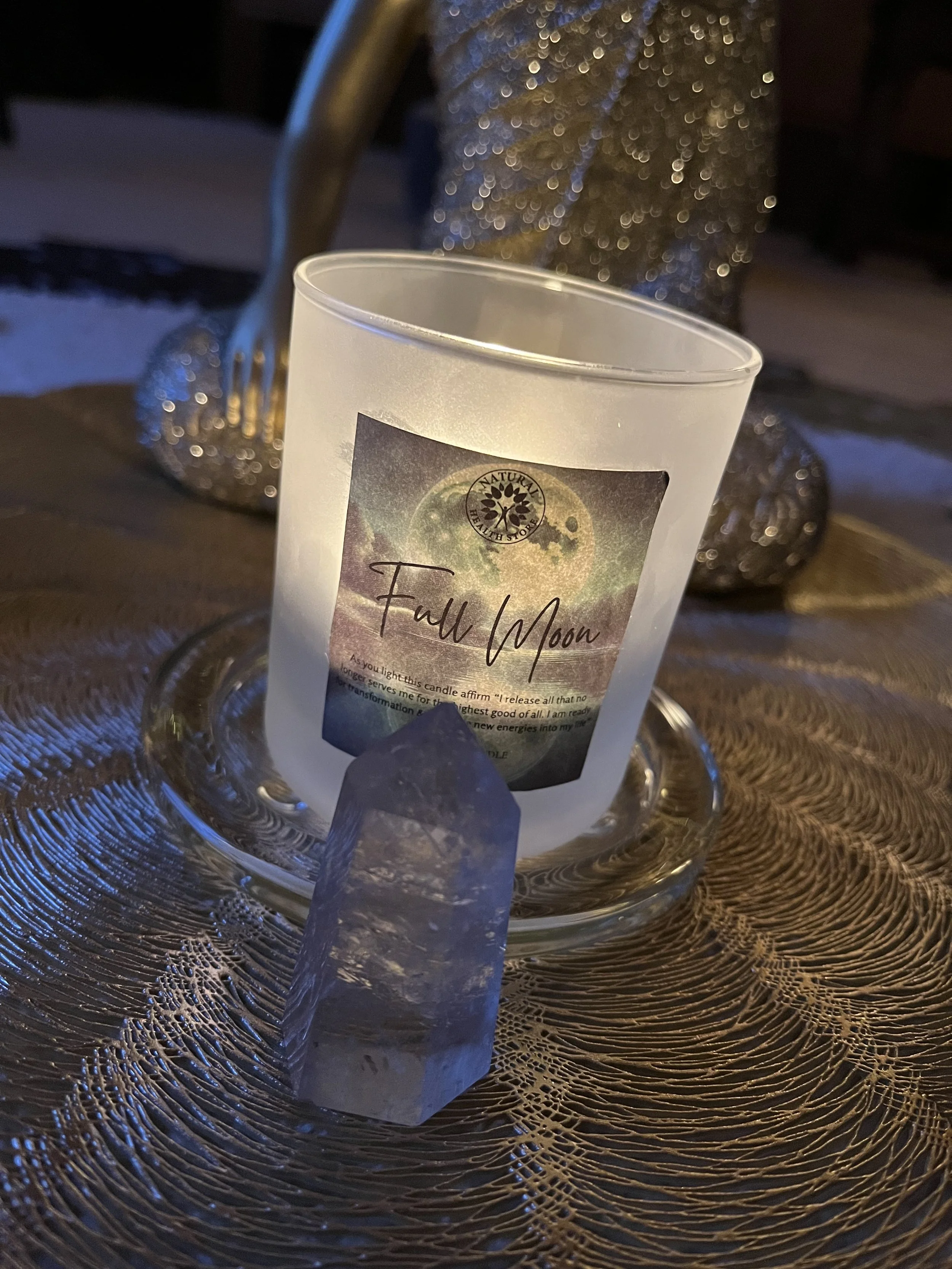 A lit candle labeled 'Full Moon' in a frosted glass holder, placed on a decorative metallic placemat with a large clear crystal in front, a metallic sculpture in the background.