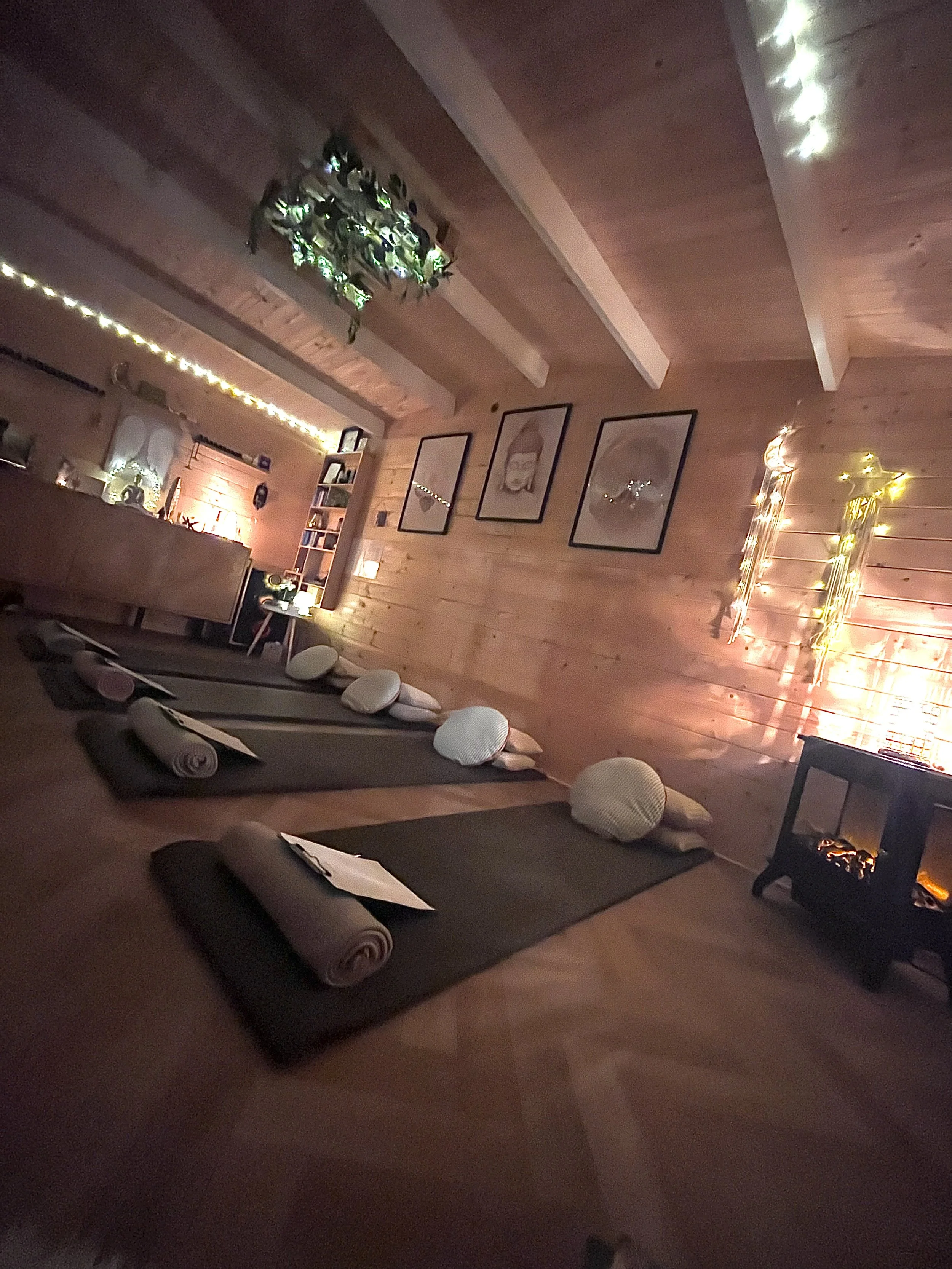 A cozy, dimly lit yoga or meditation studio with four mats laid out on the wooden floor, each accompanied by a rolled blanket and a round pillow. The walls are wooden with framed pictures, string lights, and a decorative plant hanging from the ceiling. A small table with a paper and pen is at the bottom of the mats, and a heater near the wall adds warmth to the space.