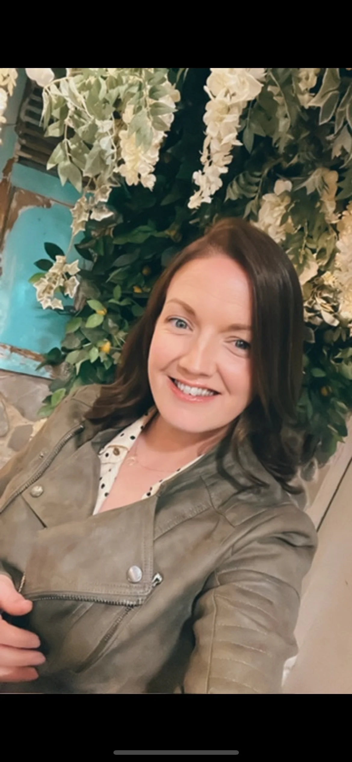 A woman with brown hair smiling, wearing a light brown jacket, standing in front of a wall of white flowers and green leaves.