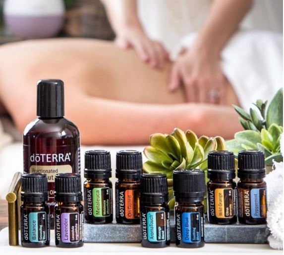 A collection of dōTERRA essential oils in front of a massage therapy scene with a person receiving a massage, and a focus on aromatic plants.