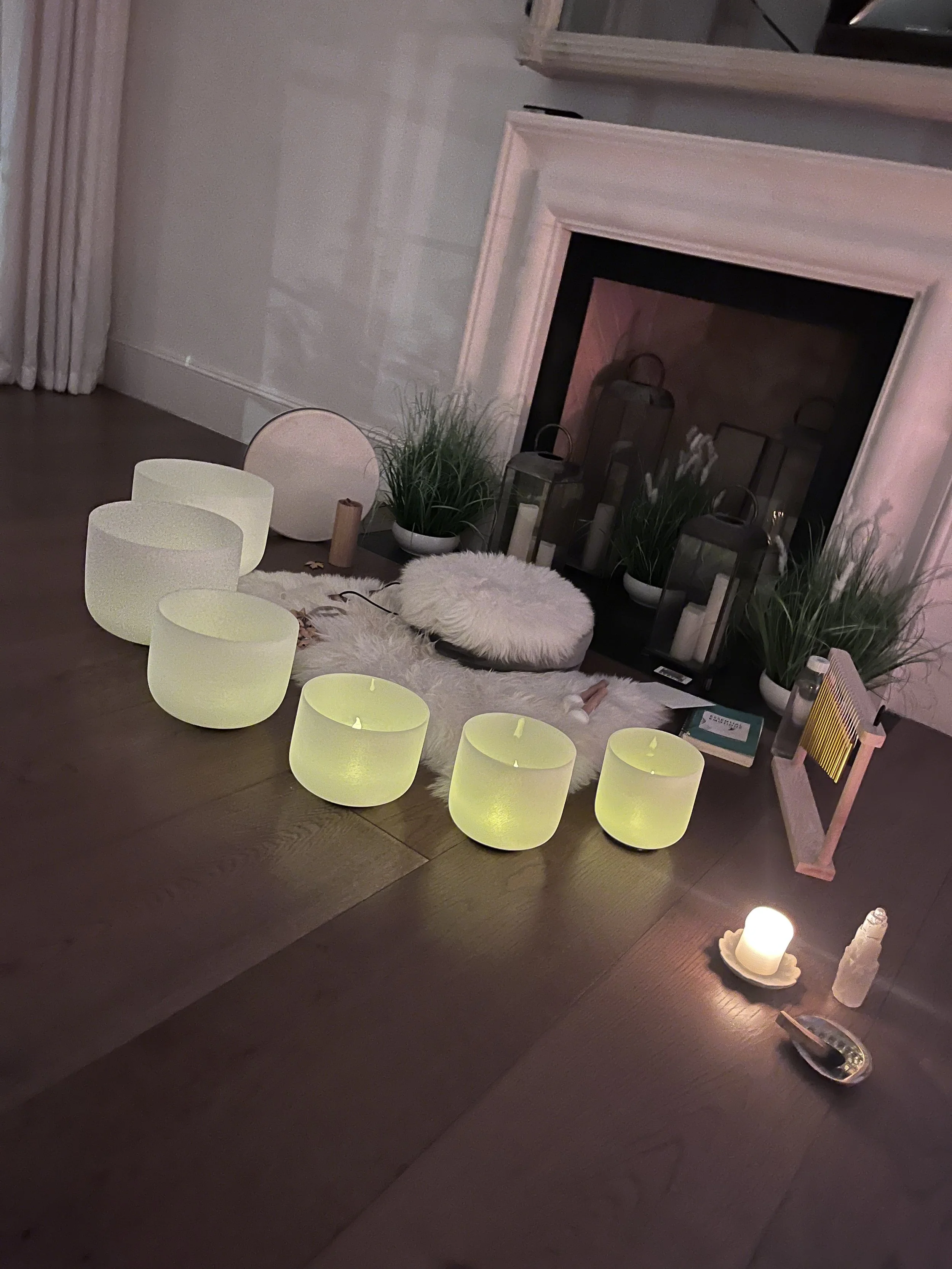 A cozy indoor space with a fire place, decorated with various candles, lanterns, potted plants, and a fluffy white seat cushion on a dark wood table.