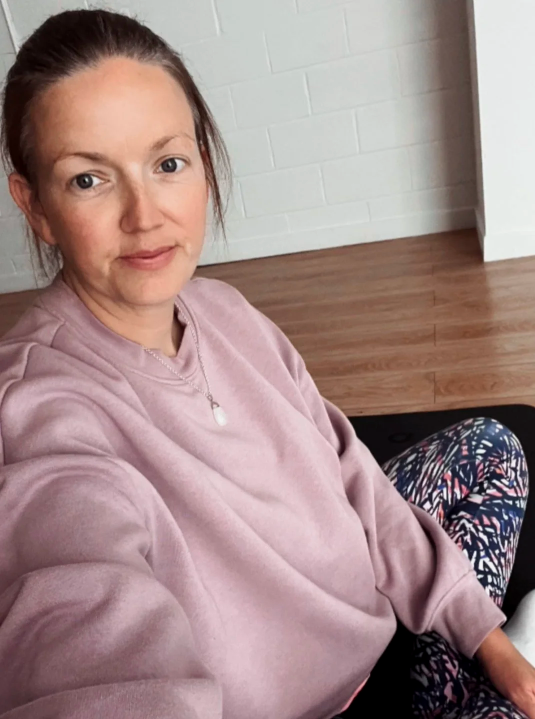 A woman taking a selfie while sitting on a black yoga mat in a room with wooden flooring and white brick walls. She has brown hair tied back, is wearing a pink sweatshirt, patterned leggings, a necklace with a pendant, and is smiling gently.