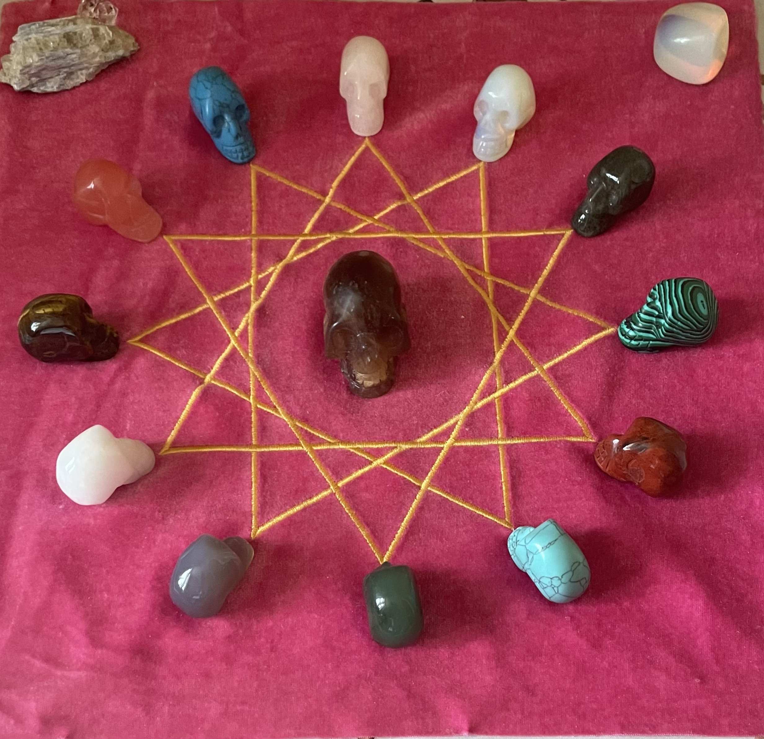 A collection of small, carved gemstone skulls arranged in a circle on a pink cloth background, with yellow string forming a geometric pattern connecting the skulls.
