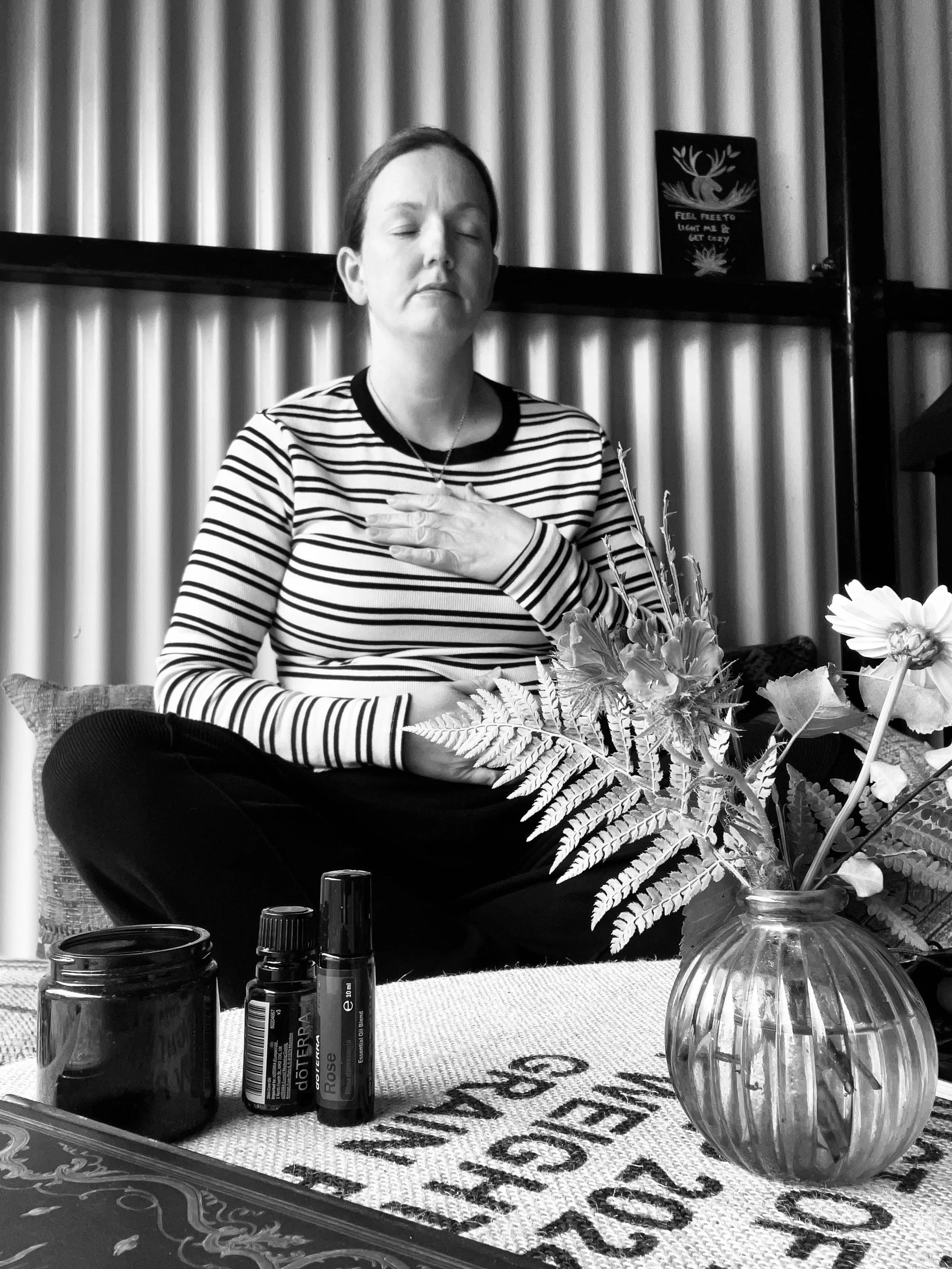 A woman sitting cross-legged on a bench with her eyes closed and hand on her chest, practicing meditation or mindfulness. In front of her, there is a table with essential oils, a flower vase, and a towel with partial text. The background features a corrugated metal wall and a small poster with a deer silhouette.