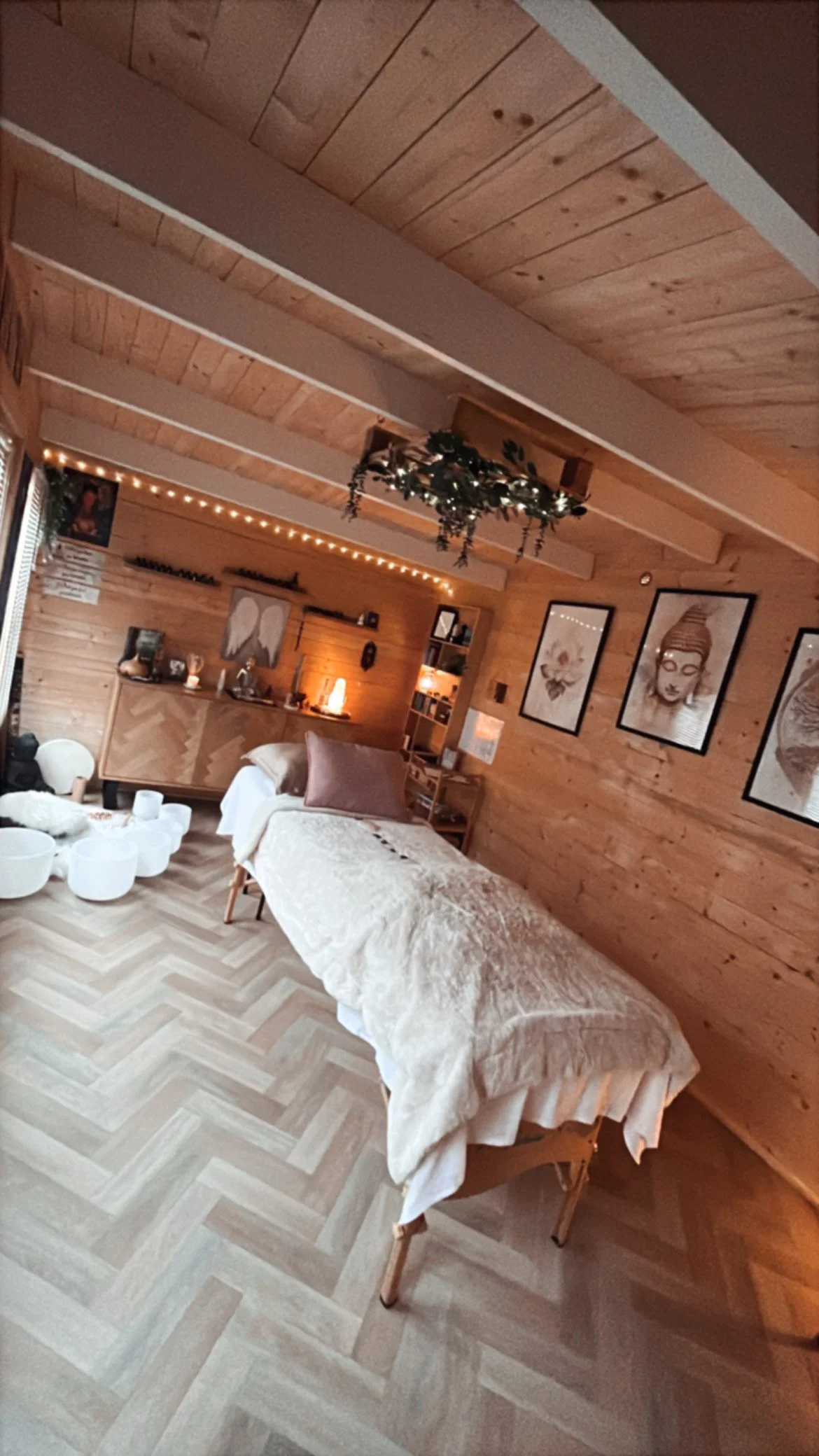A cozy wellness or massage room with wooden walls and ceiling, featuring a massage table with a beige quilted cover and a purple pillow, surrounded by wall art, fairy lights, and decorative items, creating a relaxing ambiance.