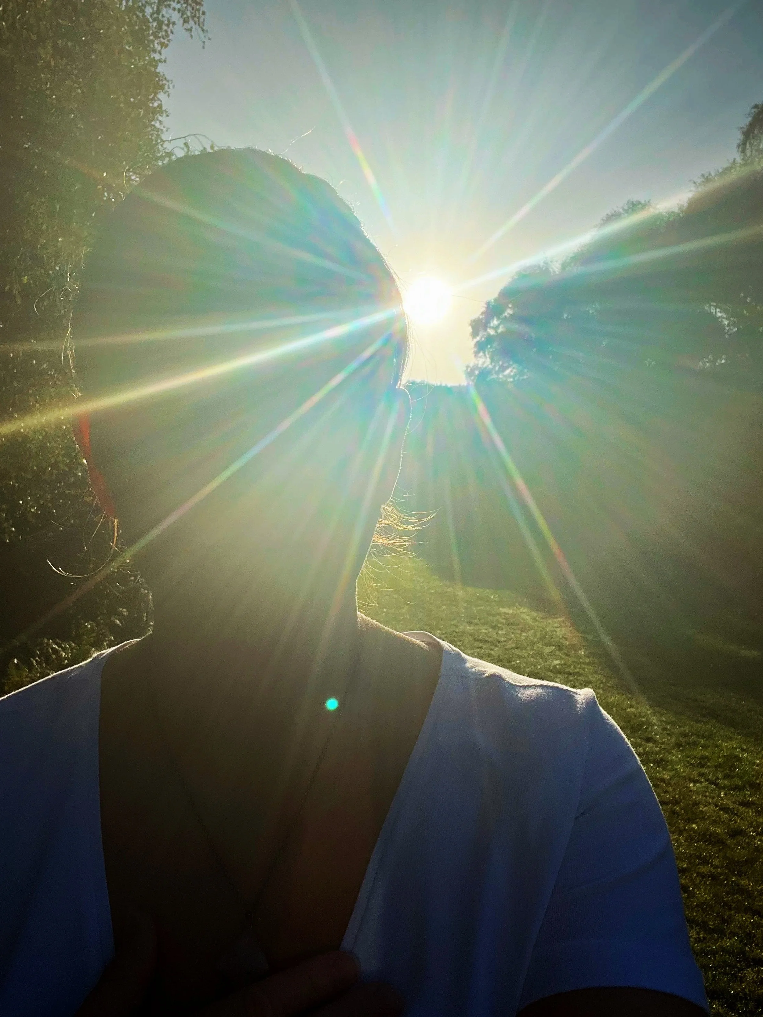 A person with their hair tied back, standing outdoors in front of the sun with sunlight creating a starburst effect. The person appears to be holding their shirt or collar with one hand. The background includes trees and grass.