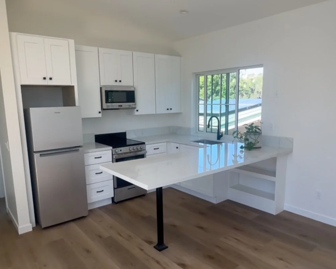 Modern kitchen with white cabinets, stainless steel appliances, and a marble countertop with an island. A window over the sink reveals a view of trees.